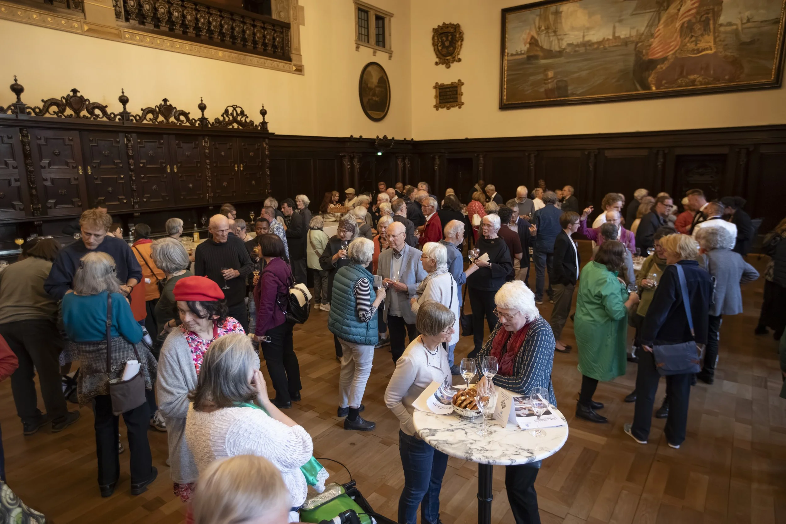 A crowded indoor event with many people socializing and tasting drinks in a large, ornate room with dark wood paneling and historical paintings on the walls.