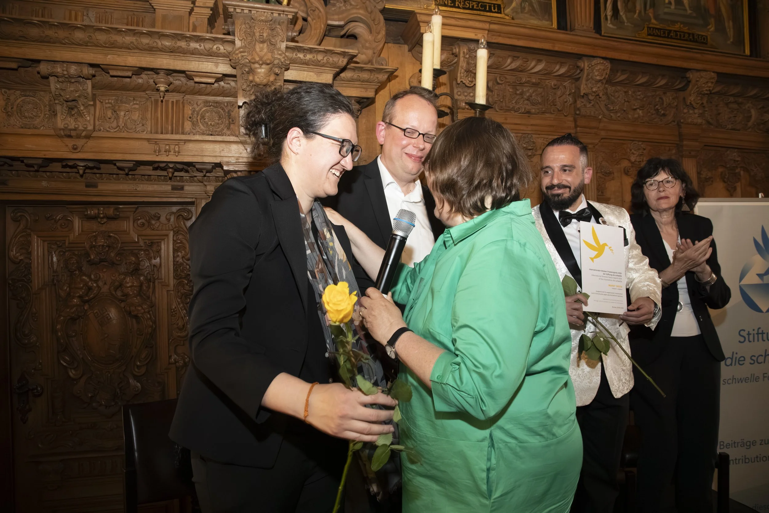 People celebrating and exchanging flowers in a formal setting with wood-carved walls and candles in the background.