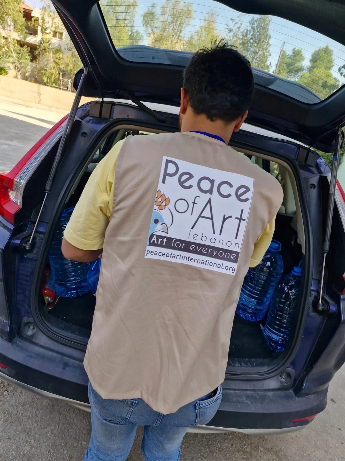 A person wearing a beige vest with a Peace of Art Lebanon logo is loading bottles of water into the back of a car, with the trunk open, in an outdoor setting with trees and buildings in the background.