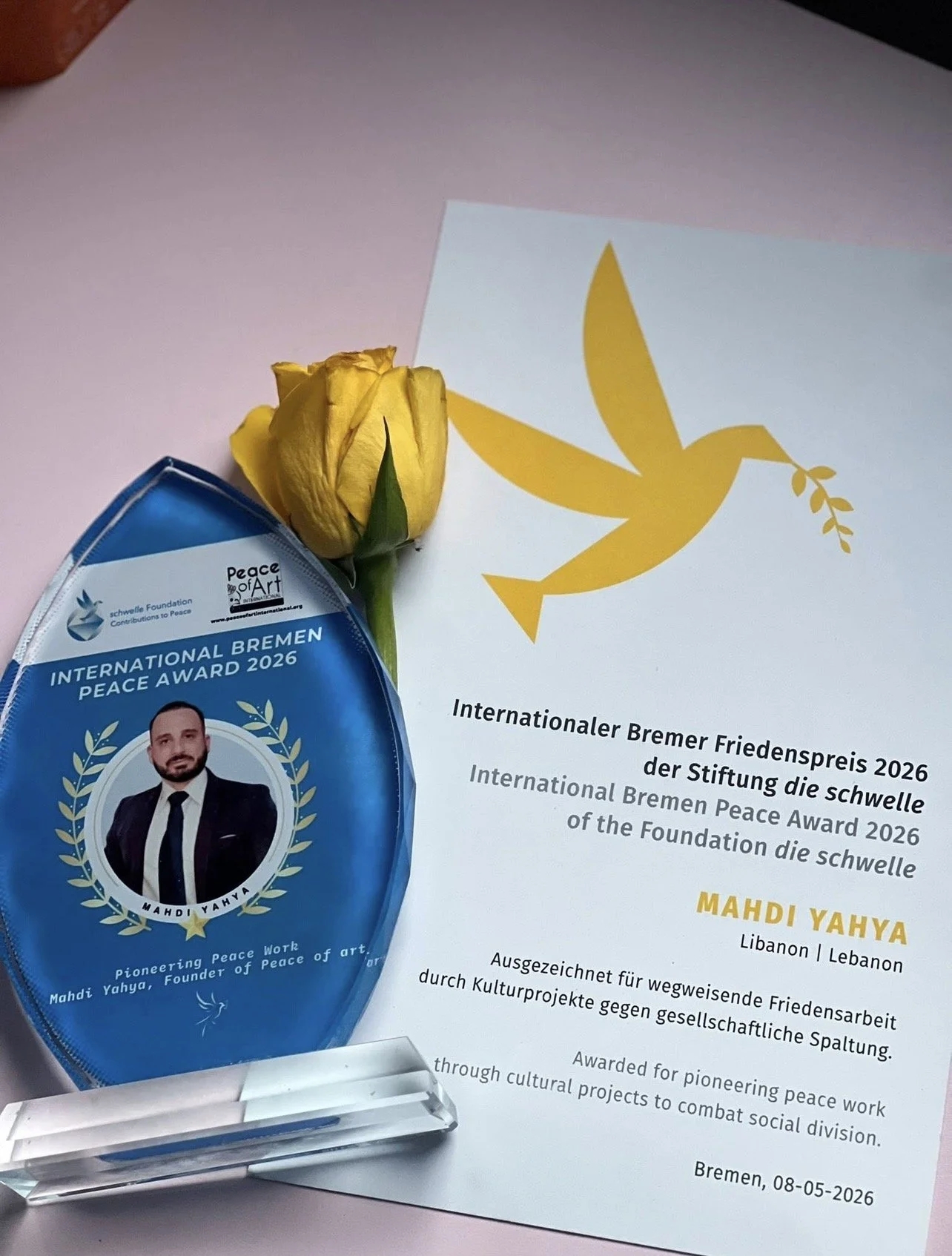 A yellow flower, a blue award badge with a man's photo and text, and a document with a yellow dove logo and text in German and English.