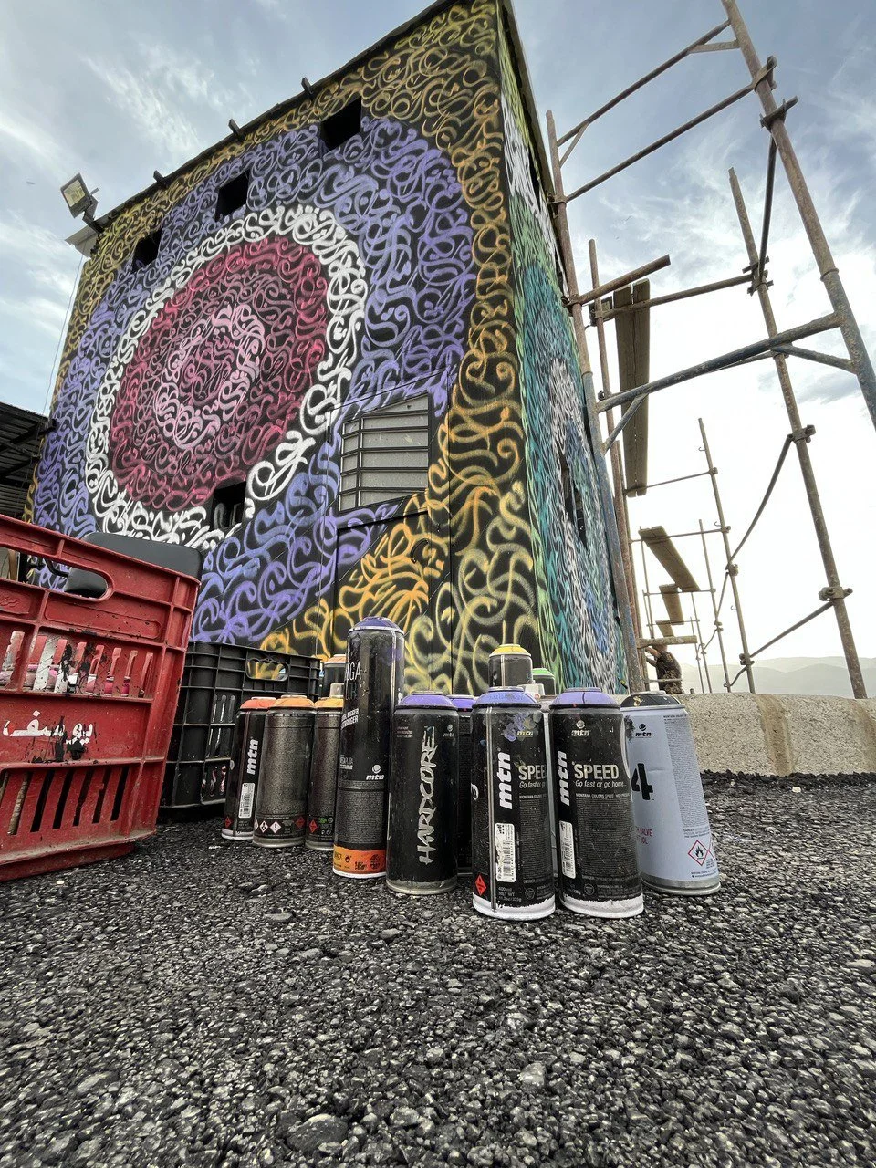 Colorful graffiti mural on a building wall, with spray paint cans and crates in the foreground, and scaffolding on the side of the building.