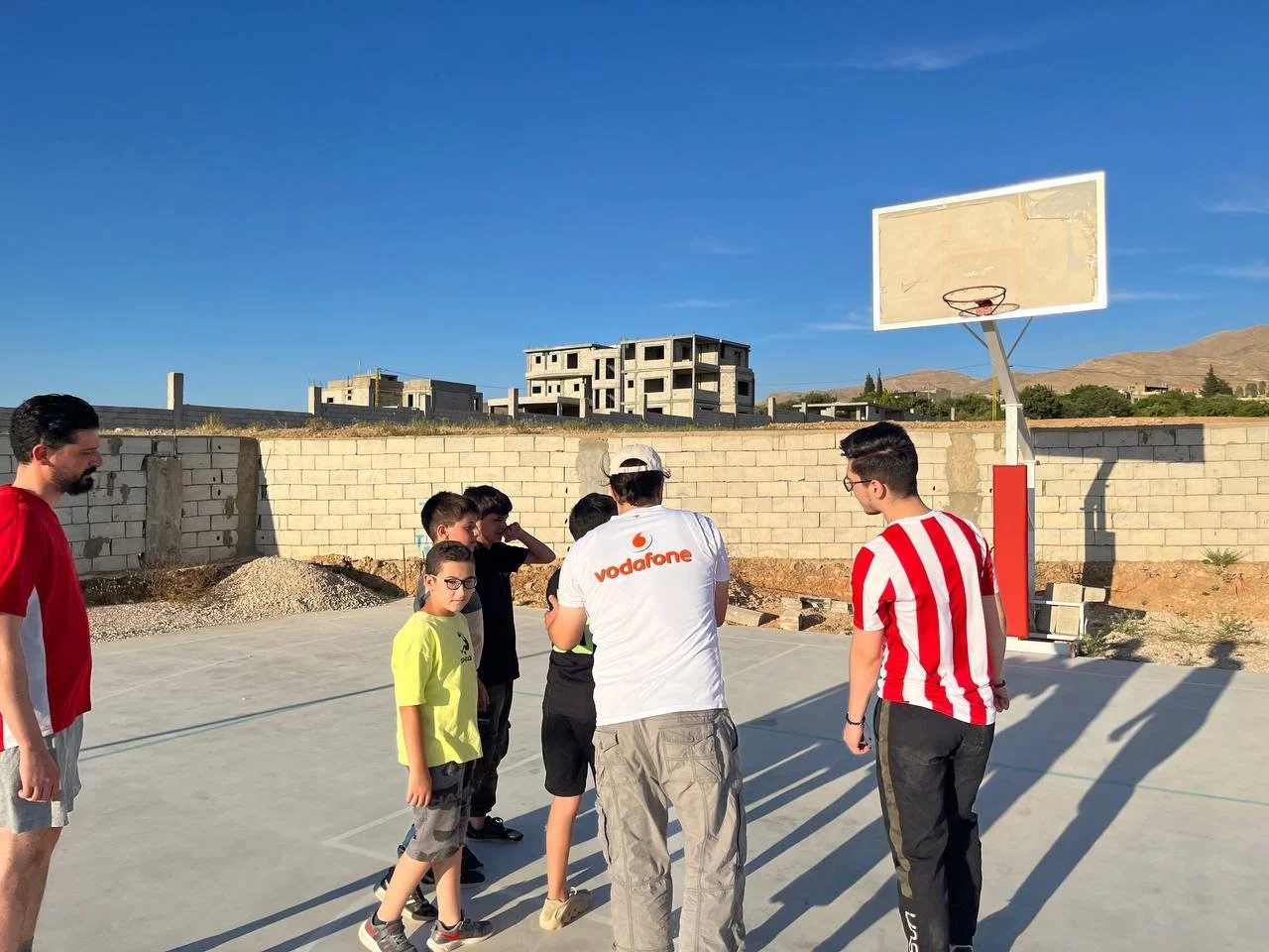 A group of children and teenagers gathered on a basketball court outdoors, with three adults, under a clear blue sky, and an unfinished building and mountains in the background.