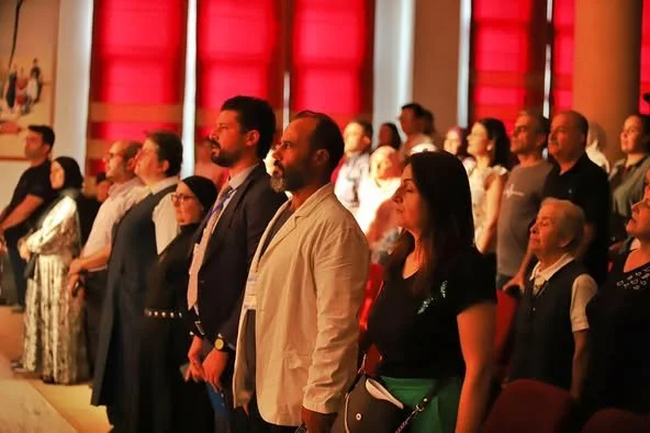 Group of people standing and facing forward in an auditorium or conference hall with red curtains in the background.