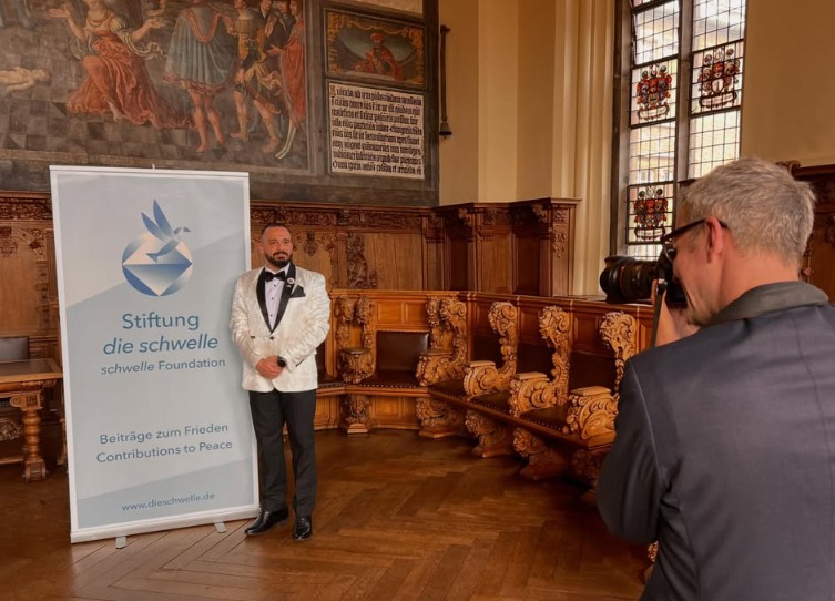 A man in a tuxedo stands beside a banner for 'Stiftung die schwelle' foundation, with wooden church pews and stained glass windows in the background, while another man in a suit photographs him.
