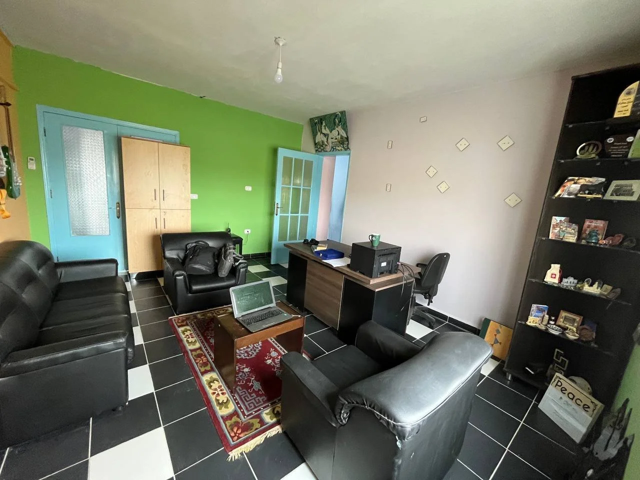 Living room with black leather furniture, green and white walls, black and white tiled floor, a desk with office supplies, a bookshelf with decorations, and a laptop.