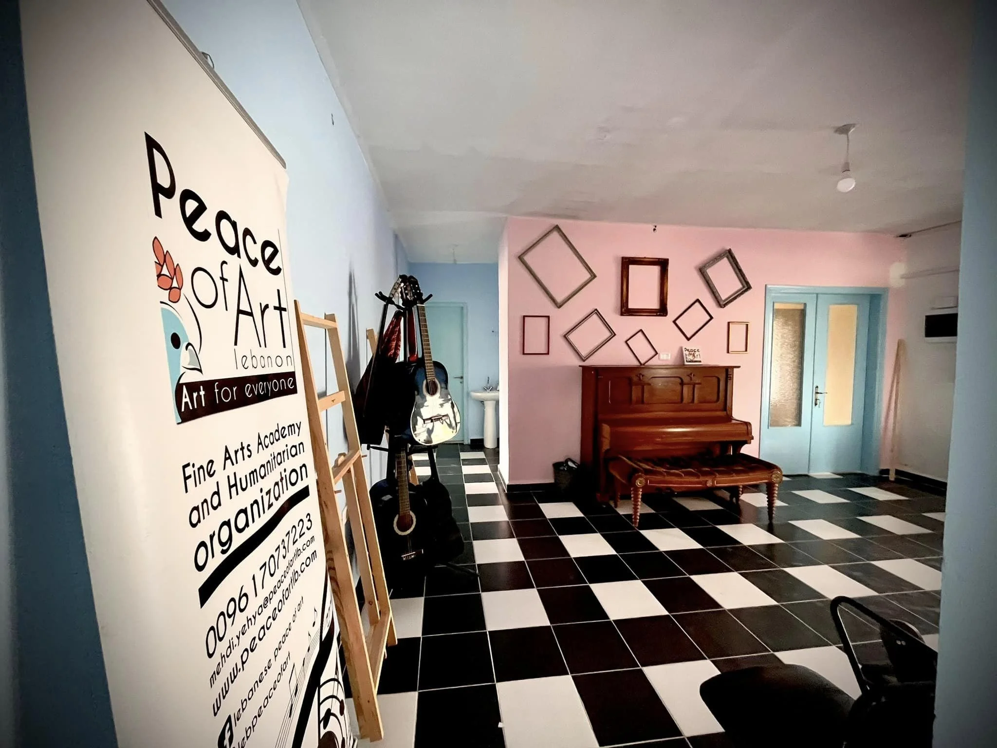Indoor room with pastel blue and pink walls, framed artwork, a wooden bench, a piano, and guitars leaning against the wall. A large sign on an easel promotes an organization, featuring a bird logo and contact information. The room has black and white