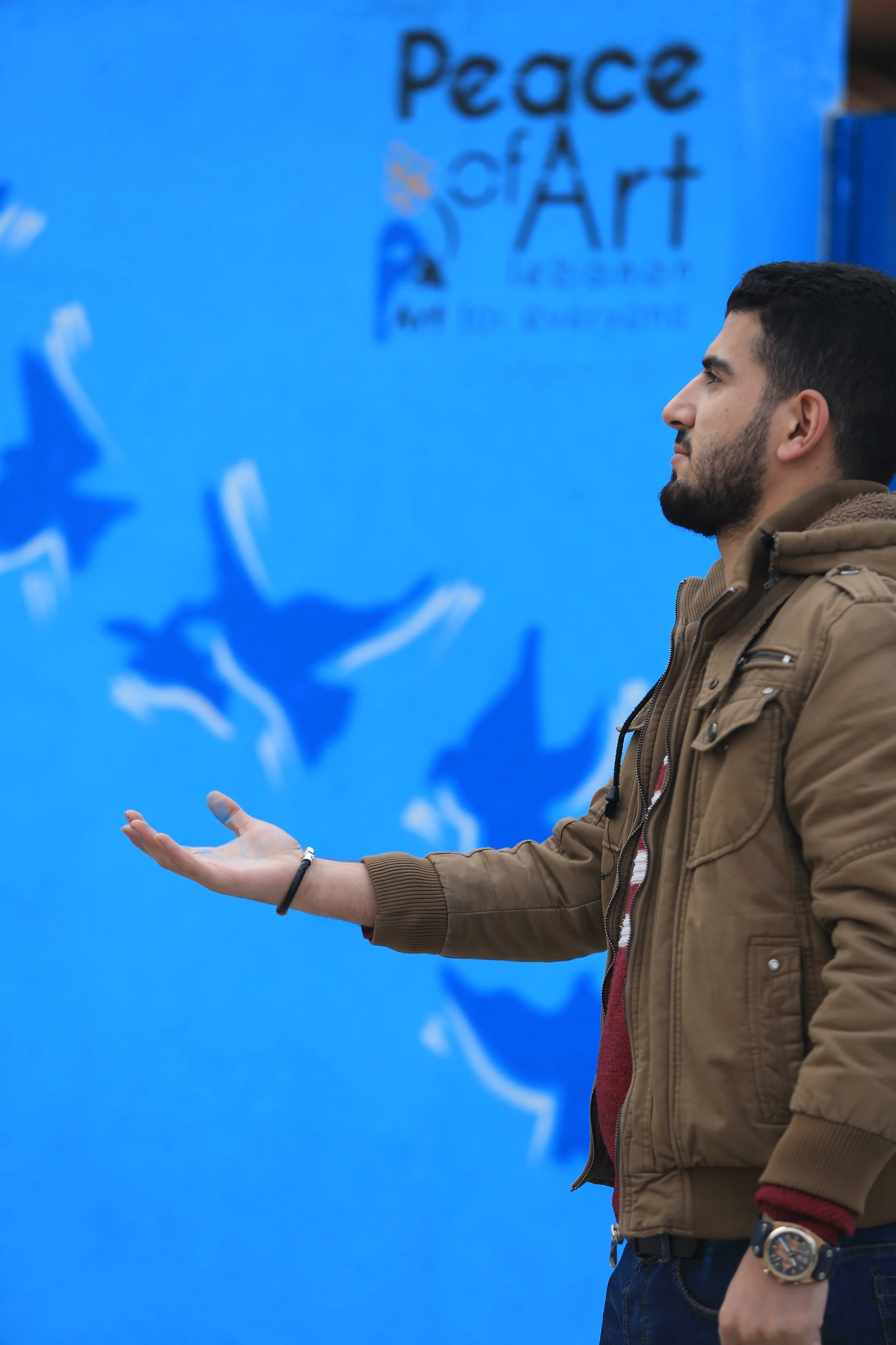 A man with a beard and short dark hair wearing a brown jacket, red sweater, and a large watch on his left wrist, standing against a blue wall with art words and symbols.
