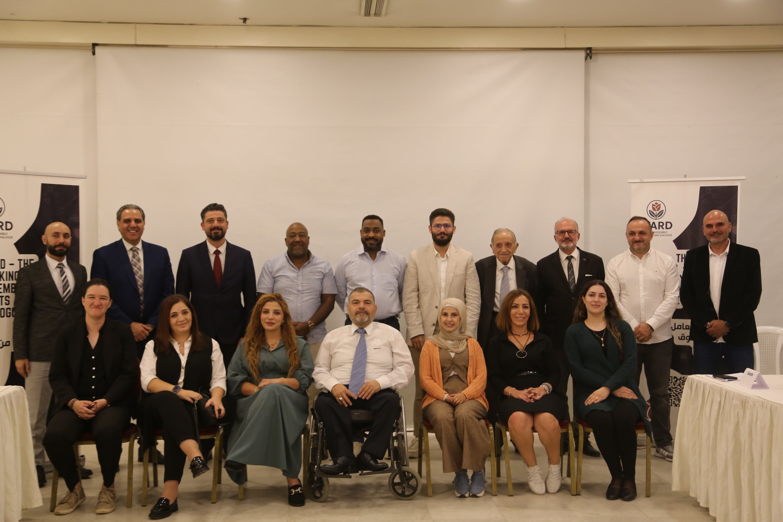 Group of diverse men and women posing for a photo at a formal event, with some seated and others standing behind. Some people are dressed in business attire, and there are banners in the background.