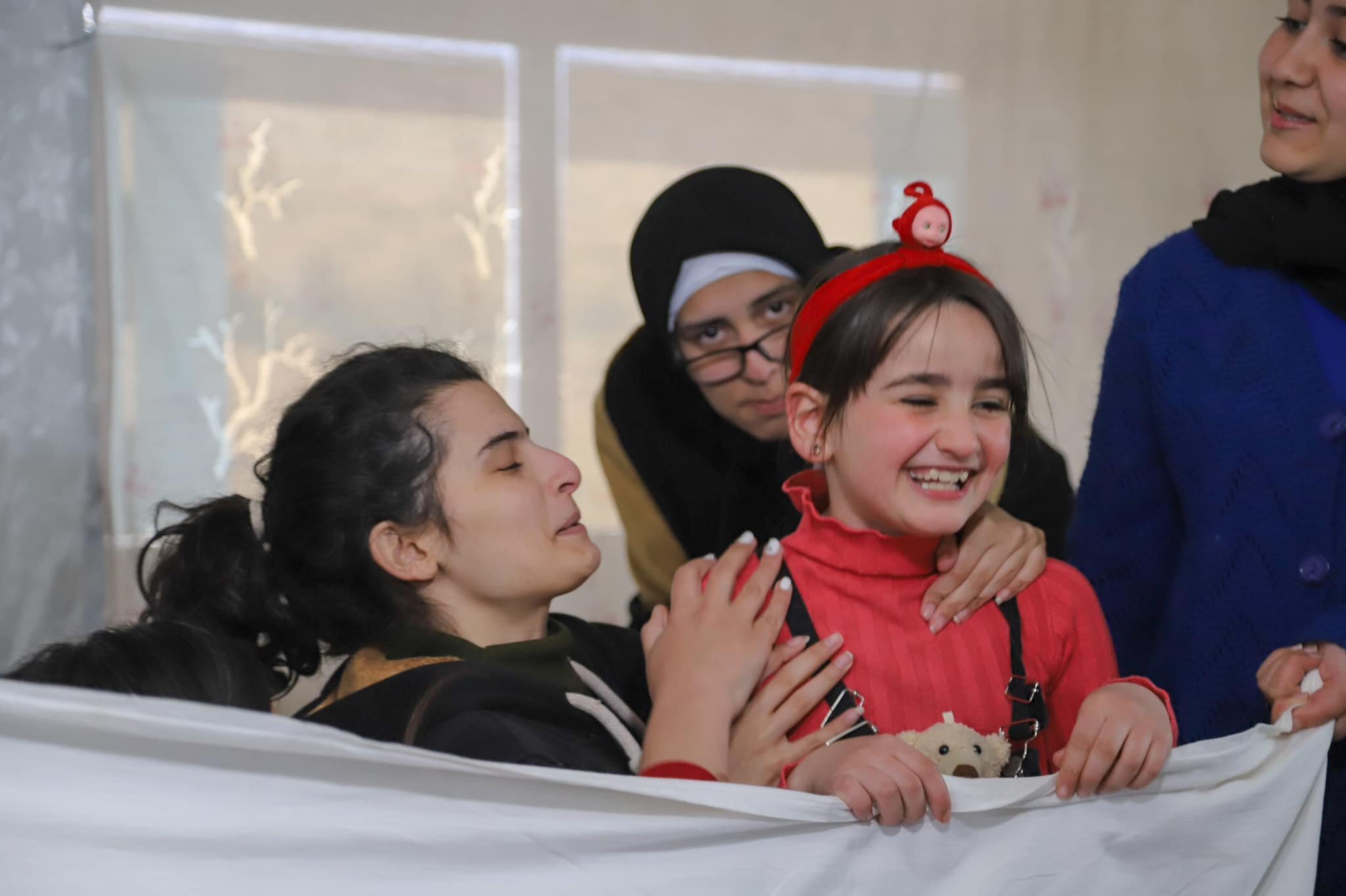Group of cheerful children and adults gathered around a blanket, with some children wearing Christmas-themed headbands, engaged in a festive activity.
