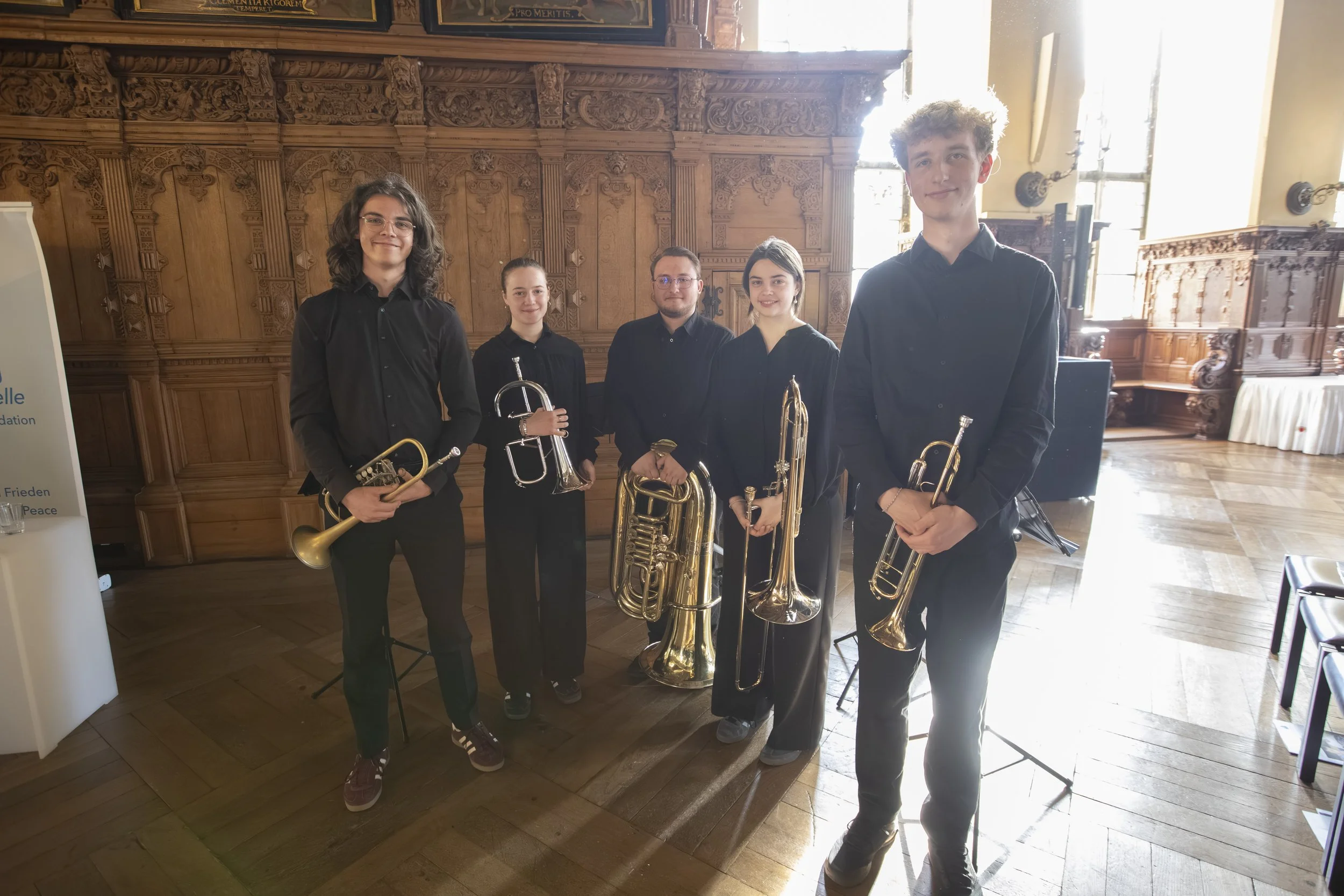 A group of five young musicians in black clothing holding brass instruments, standing in a room with ornate wood paneling and large windows.