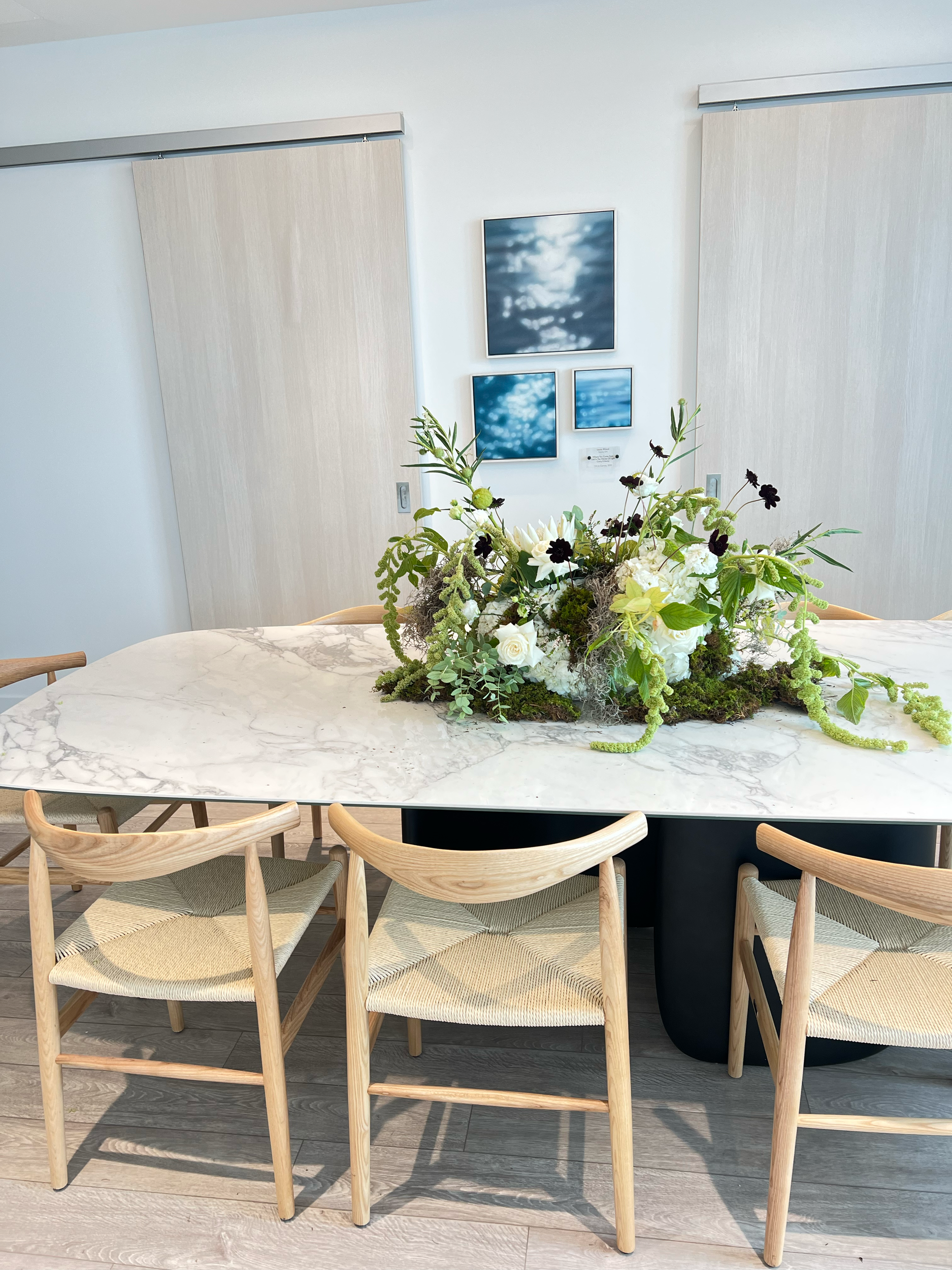 A modern marble dining table with a floral centerpiece in a bright, minimalist room with light wood chairs and abstract artwork on the wall.