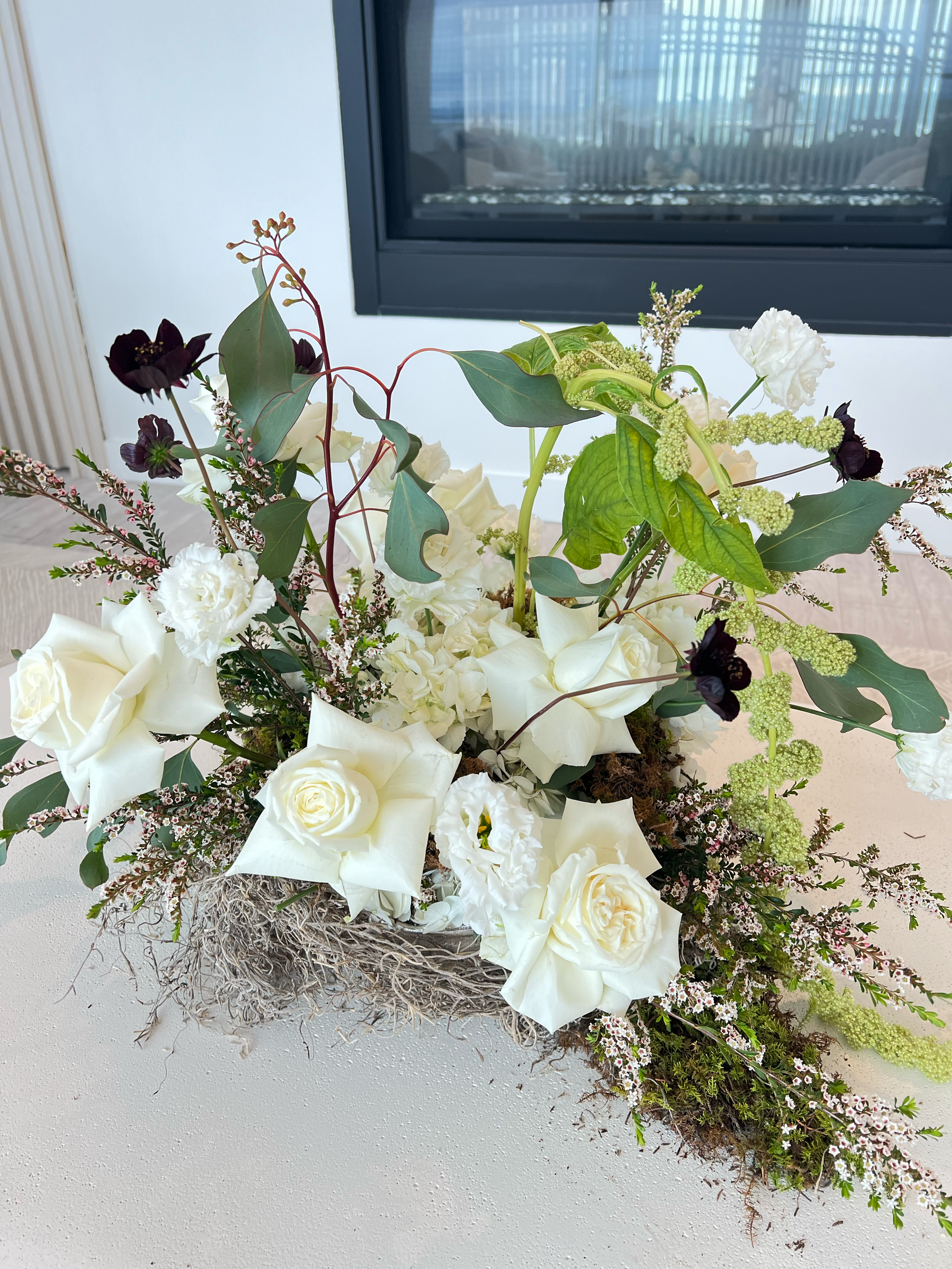 A floral arrangement in a rectangular container featuring white roses, white calla lilies, dark purple flowers, various green leaves, and small pink and white flowers, with a fire place in the background.
