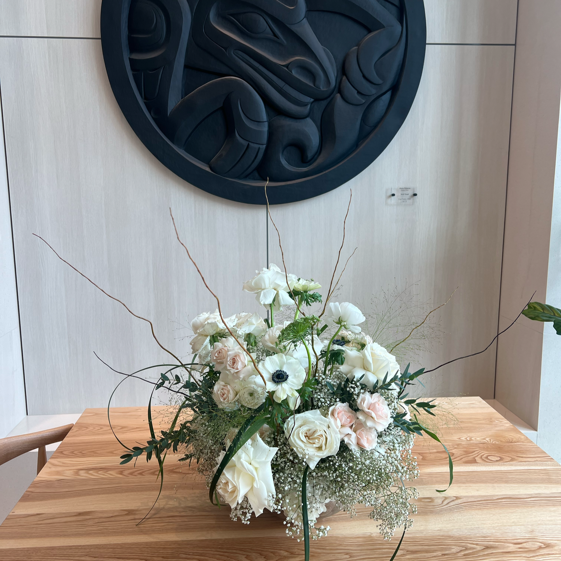 A floral arrangement of white roses, anemones, and baby's breath with greenery on a wooden table, with a black carved totem wall art above.