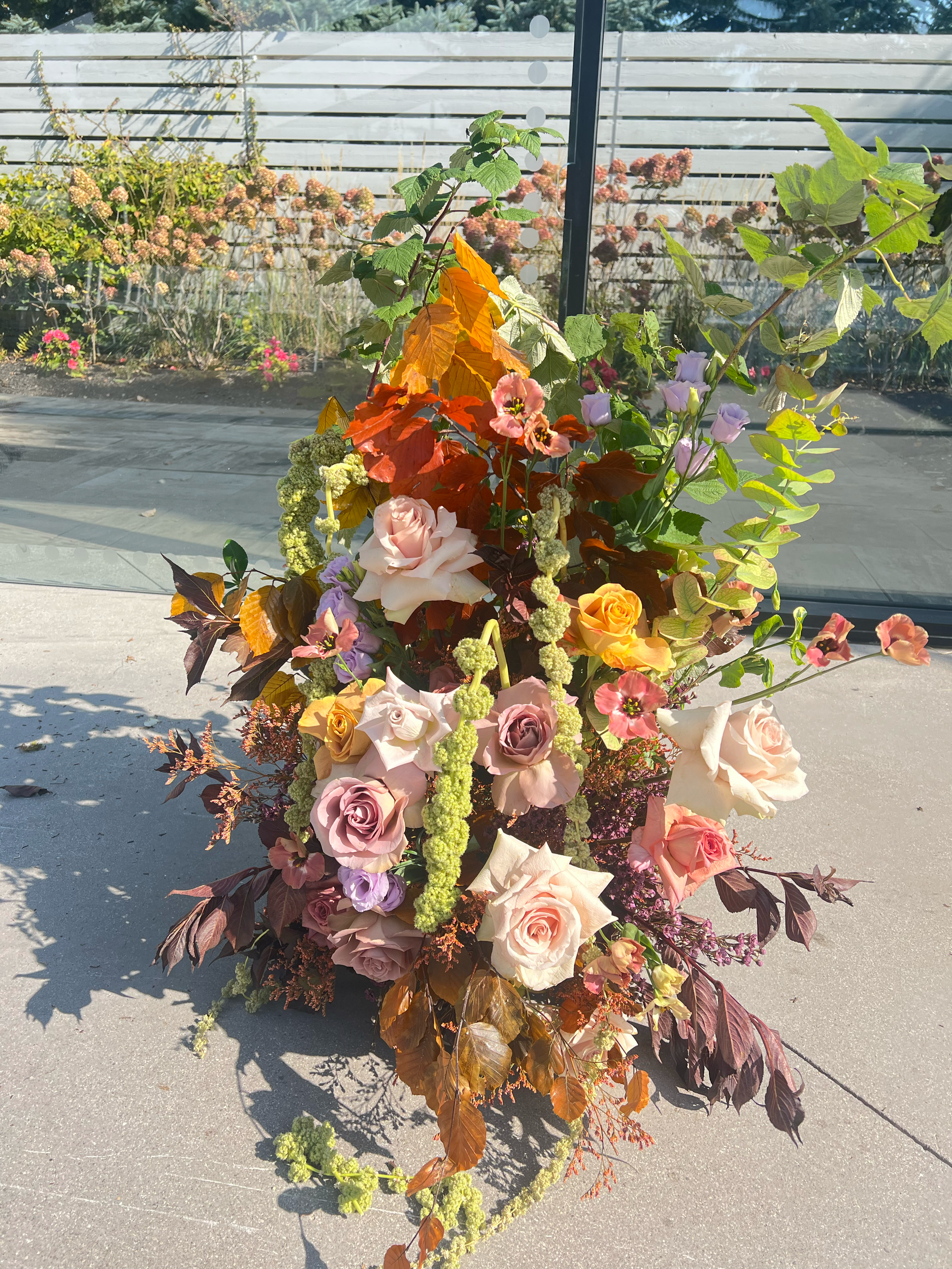 A colorful floral arrangement featuring roses, lilies, and various greenery placed outdoors on a concrete surface with a fence and garden in the background.