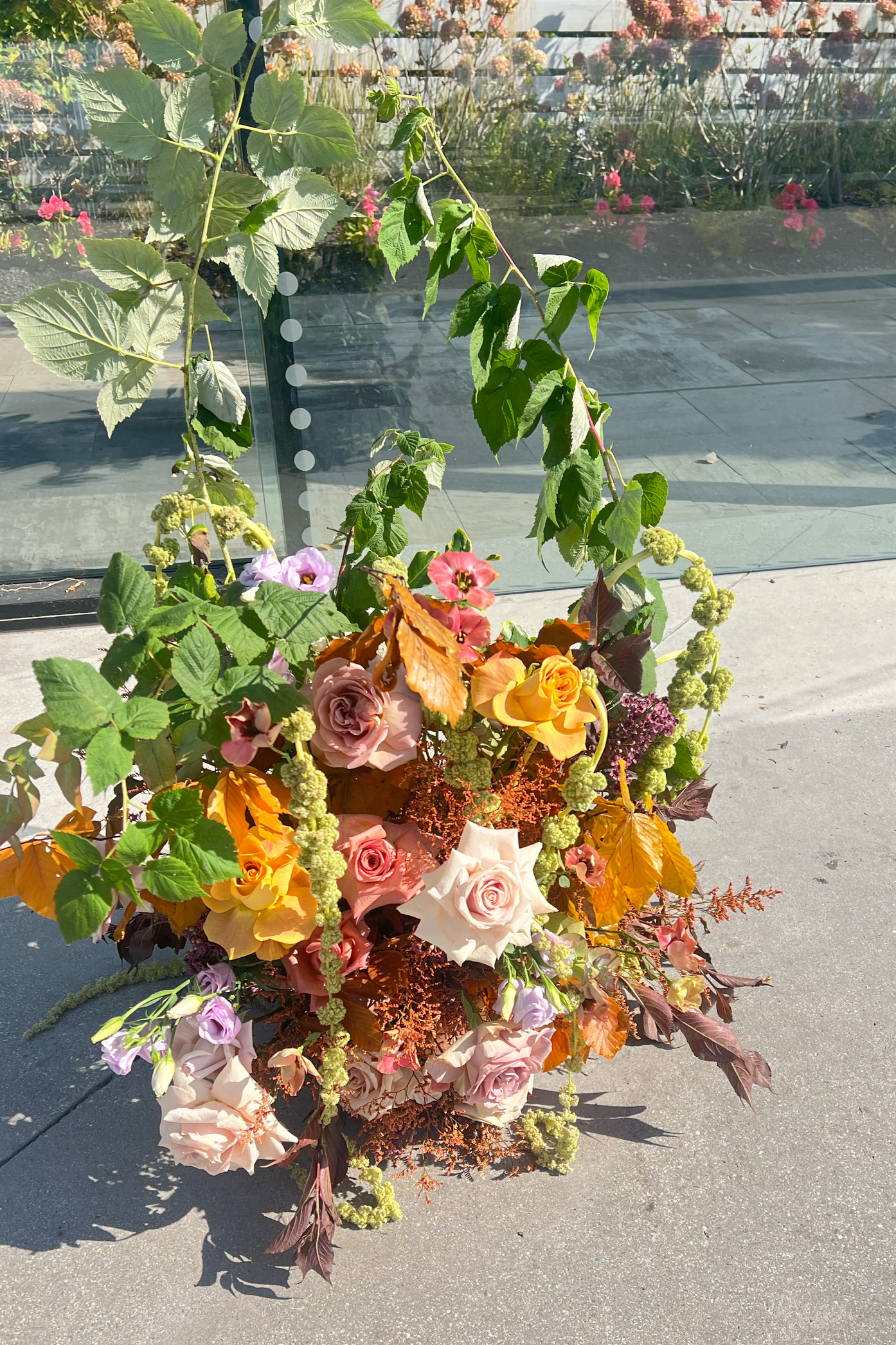 A colorful floral arrangement with roses, greenery, and other flowers, set on a concrete surface outside a glass building with flowers visible inside.