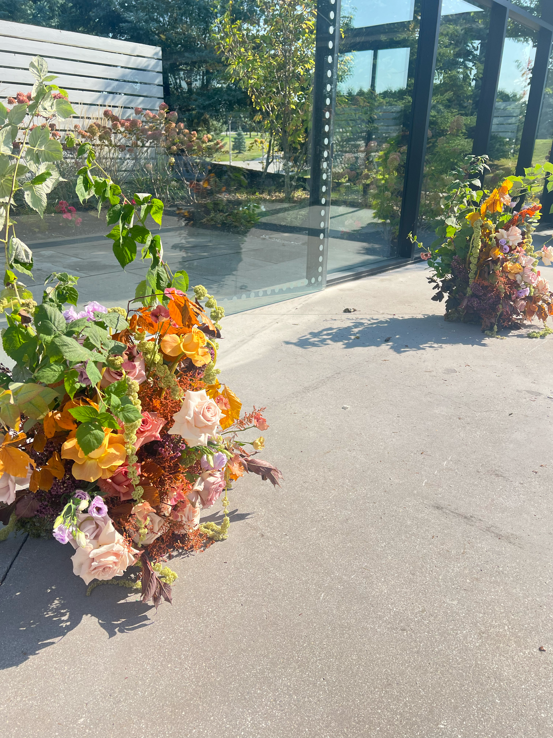 Two large floral arrangements with various colorful flowers placed outside near glass walls, with a sidewalk and greenery in the background.