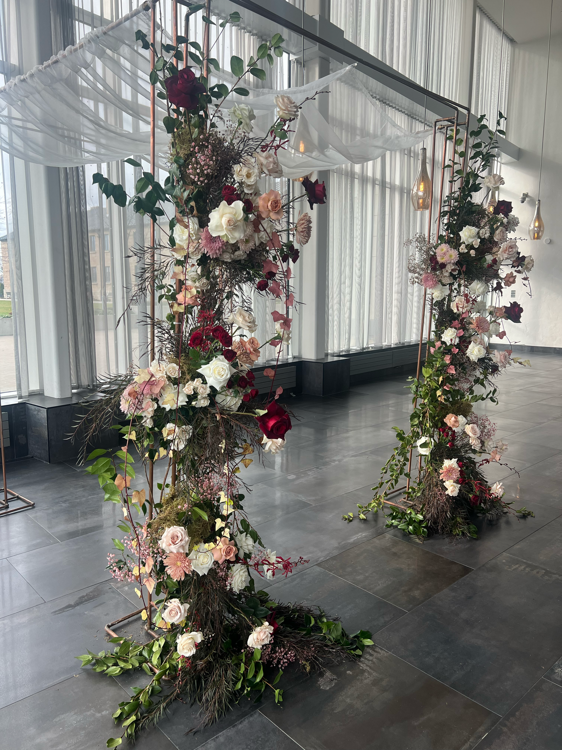 Elegant floral arch decoration with pink, white, and deep red flowers, greenery, and hanging light bulbs, set in a modern indoor space with large windows and sheer curtains.