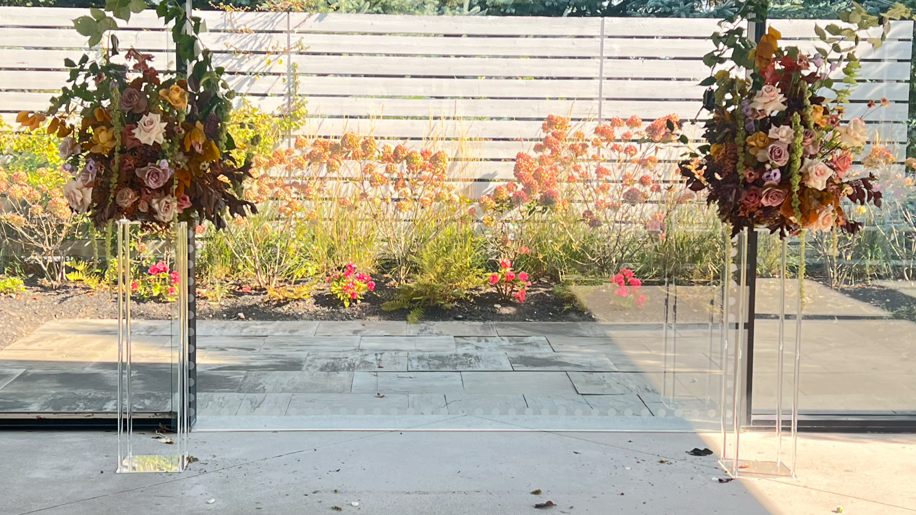Two tall flower arrangements on clear acrylic pedestals in front of a glass wall with a garden view, including pink, purple, and white roses and greenery.