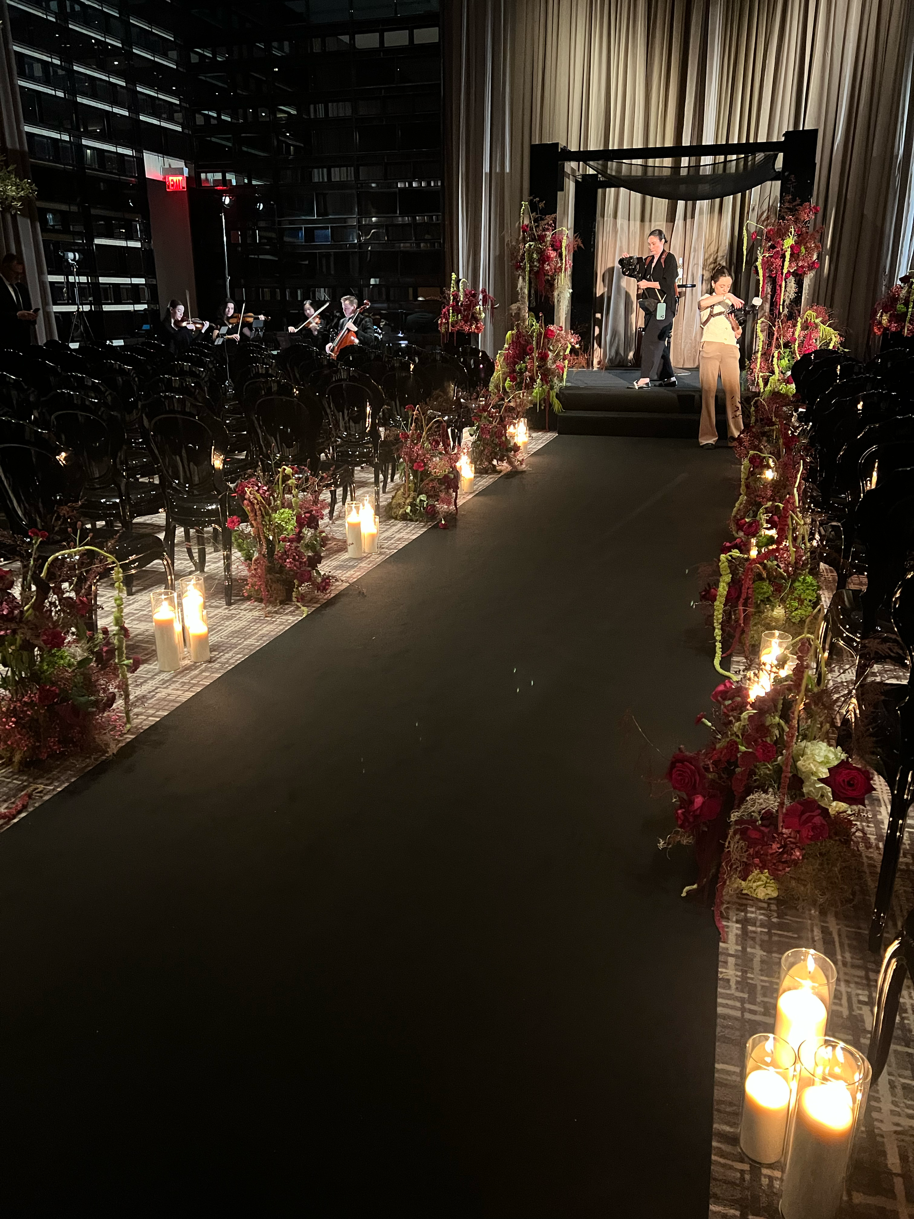 An event setup in a modern building with a stage, floral arrangements, candles, and chairs. Two people are on stage preparing or speaking, while others are seated or standing around the event space.