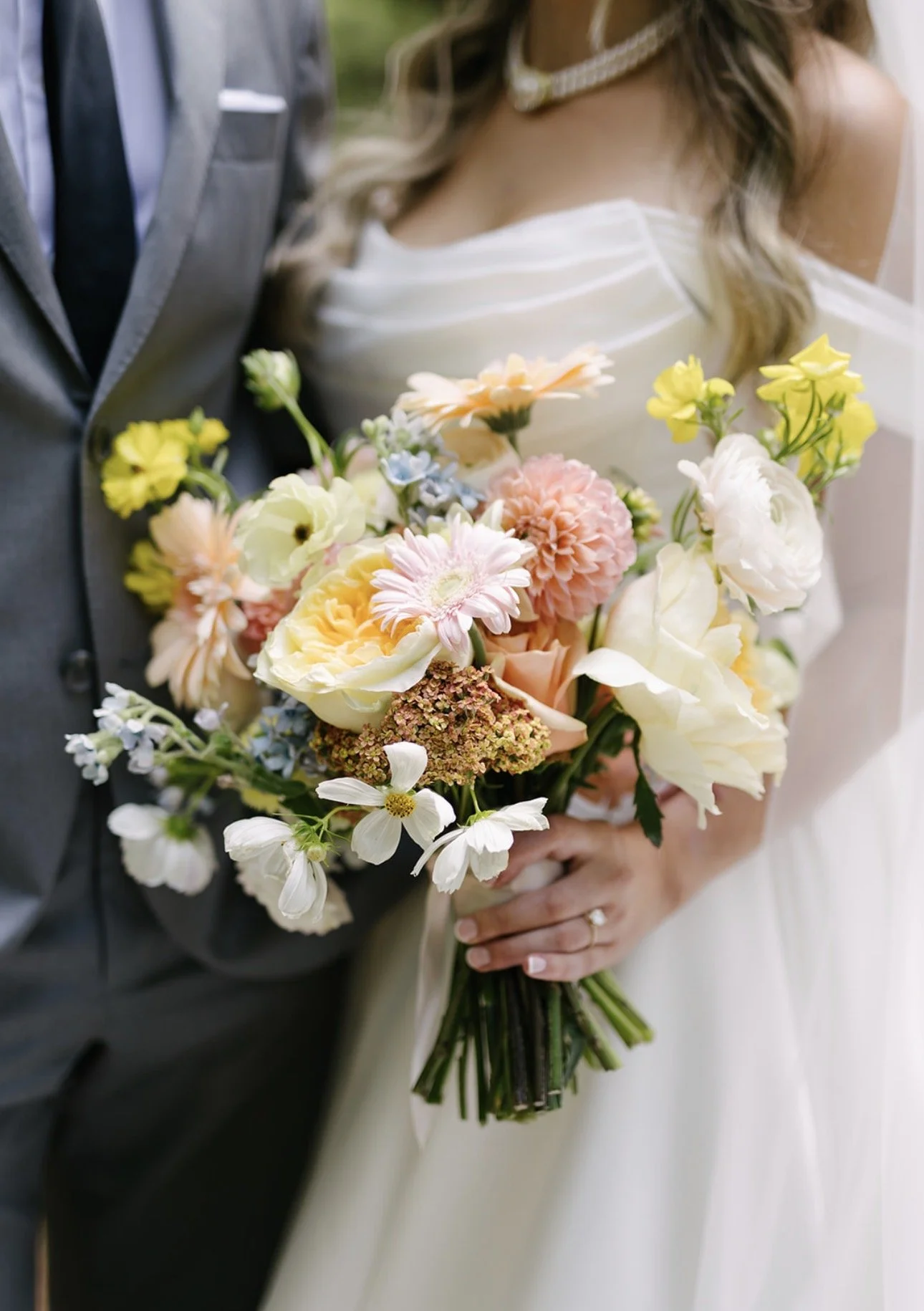 Close-up of a bride holding a colorful bouquet of flowers at her wedding, with part of a groom in a gray suit visible beside her.