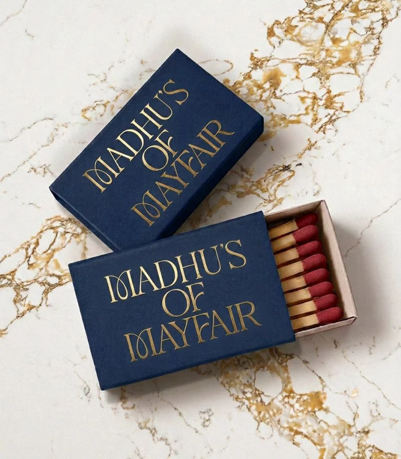 Blue match boxes with gold foil block Madhu's of Mayfair logo on a white marble table top