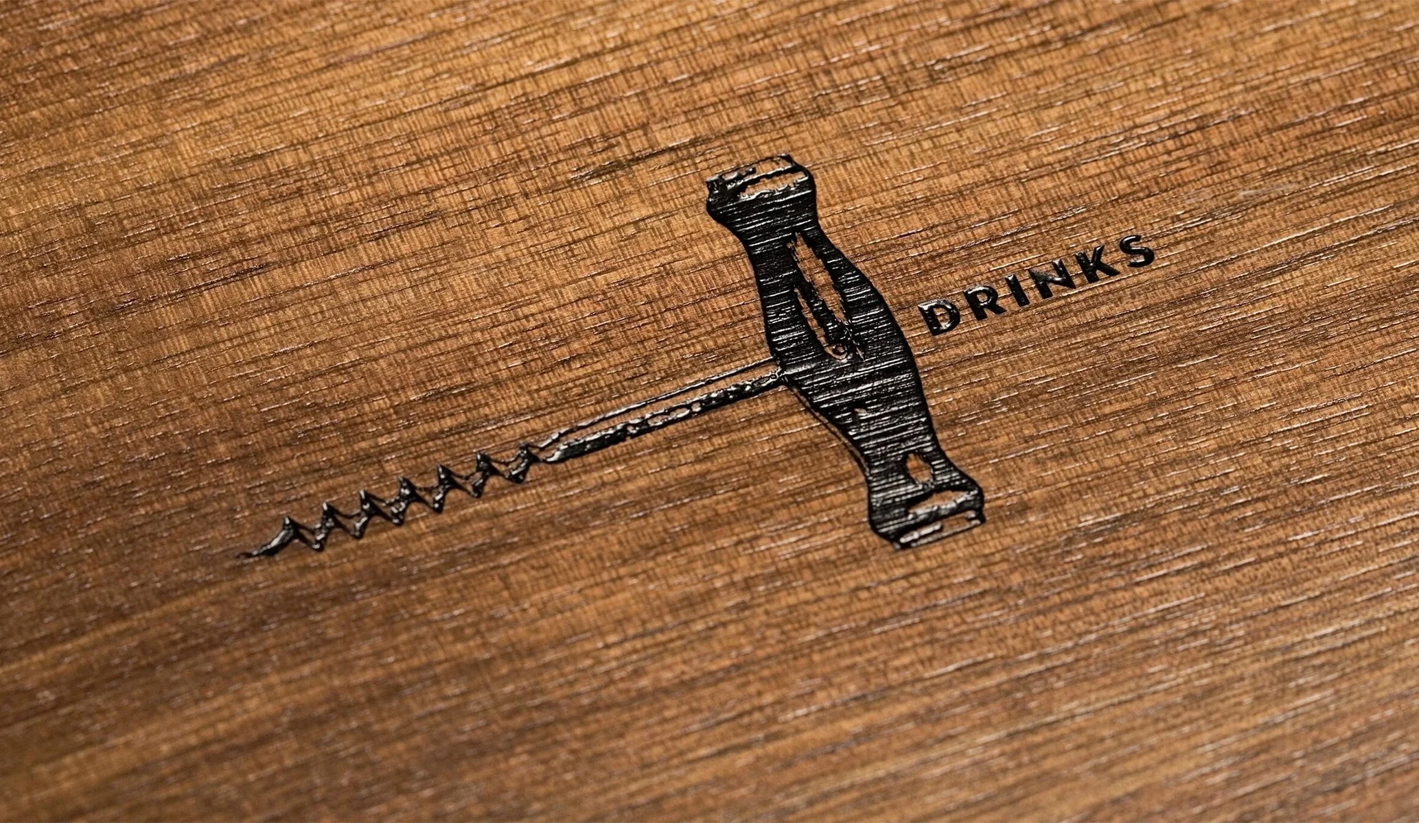 Image of a corkscrew engraved on wood, with the word 'DRINKS' next to it.