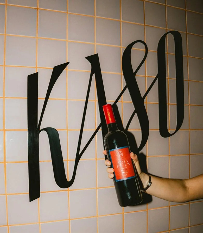 A bottle of wine being held in front of vinyl wall signage of the logo for Kaso bar in Shoreditch