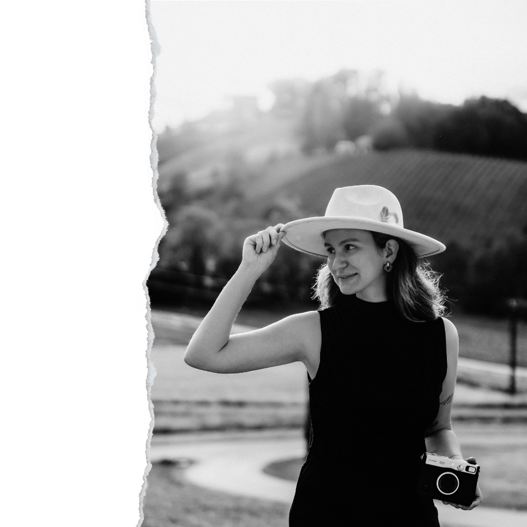 Juliana, die Fotografin, hält ihre Kamera in der Hand und blickt mit einem Lächeln in die Ferne. Sie trägt einen hellen Fedora-Hut, hinter ihr ist die Steirische Weinstraße.
