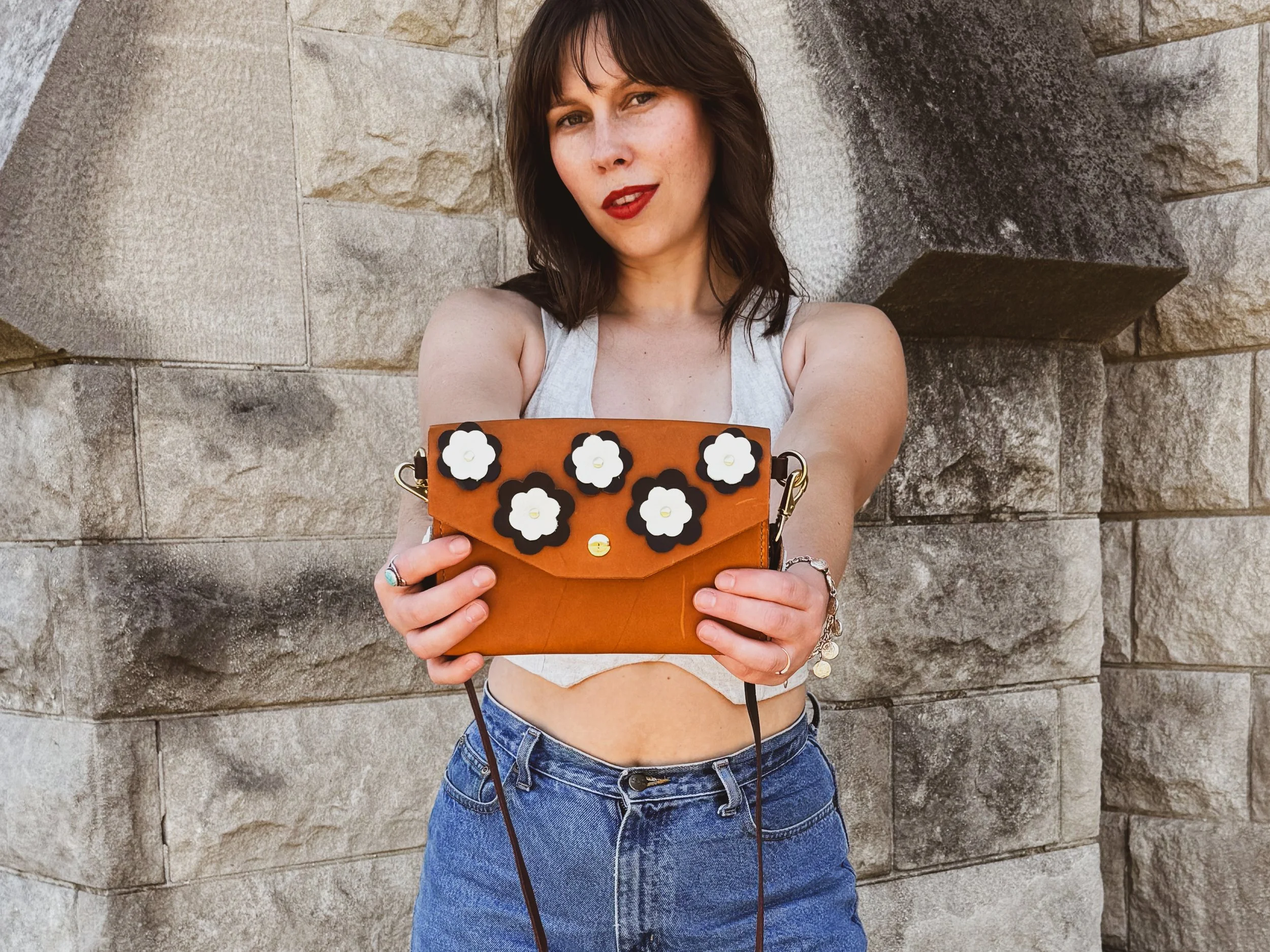 Woman with dark brown hair and red lipstick holding a brown handbag with black and white floral decorations in front of a stone wall.