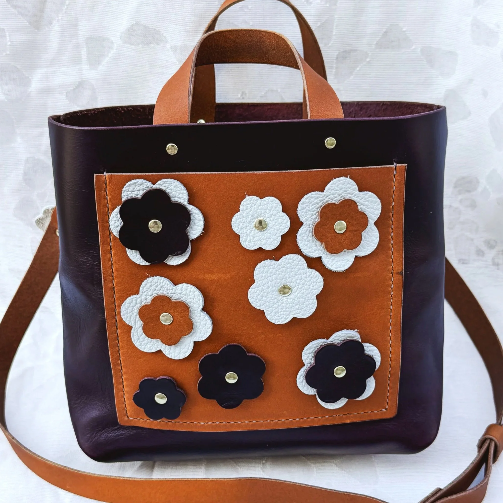 A brown and black leather handbag with a front pocket decorated with white, black, and brown leather flower appliqués.