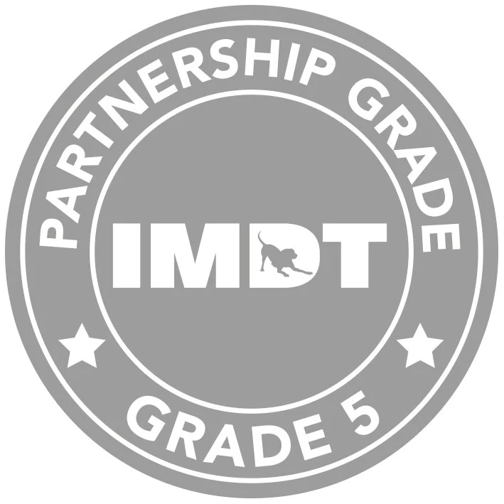 A grayscale circular badge with the text ‘PARTNERSHIP GRADE 5’ around the outer edge. The center features the letters ‘IMDT’ with a silhouette of a dog in the letter ‘D’.