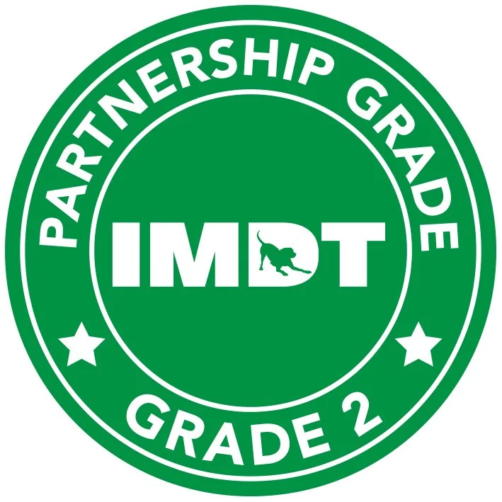 Green circular badge with white text that reads "Partnership Grade 2" around the outer edge. Inside, the center of the badge displays the letters "IMPACT," with a silhouette of a dog standing on the letter "I."