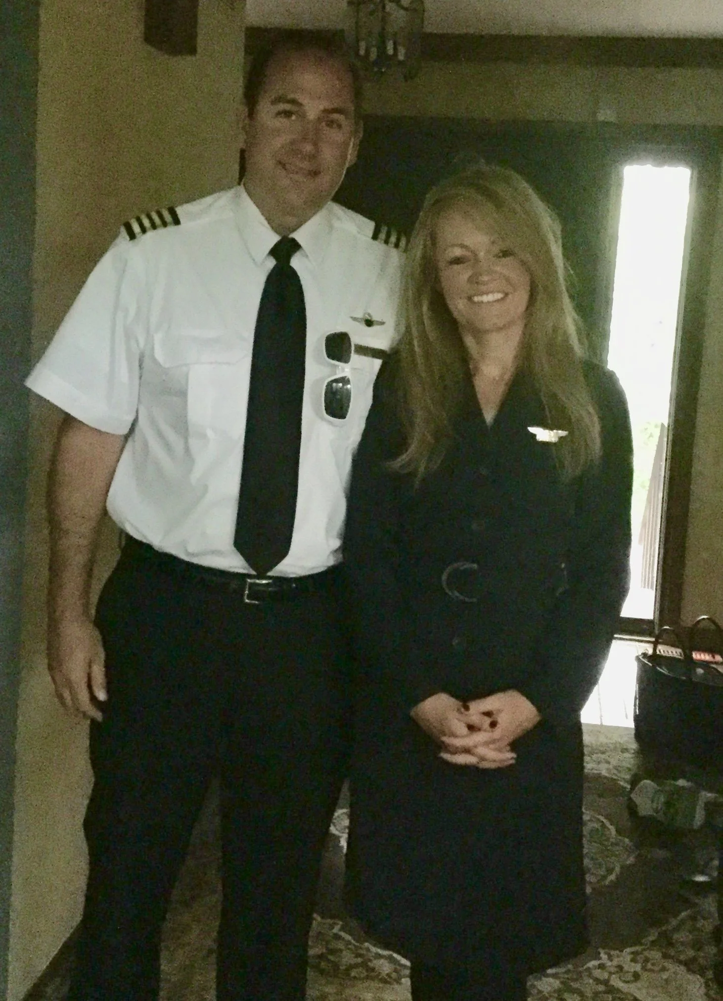 A man in a white pilot uniform and a woman in a black pilot uniform standing together indoors, both smiling at the camera.