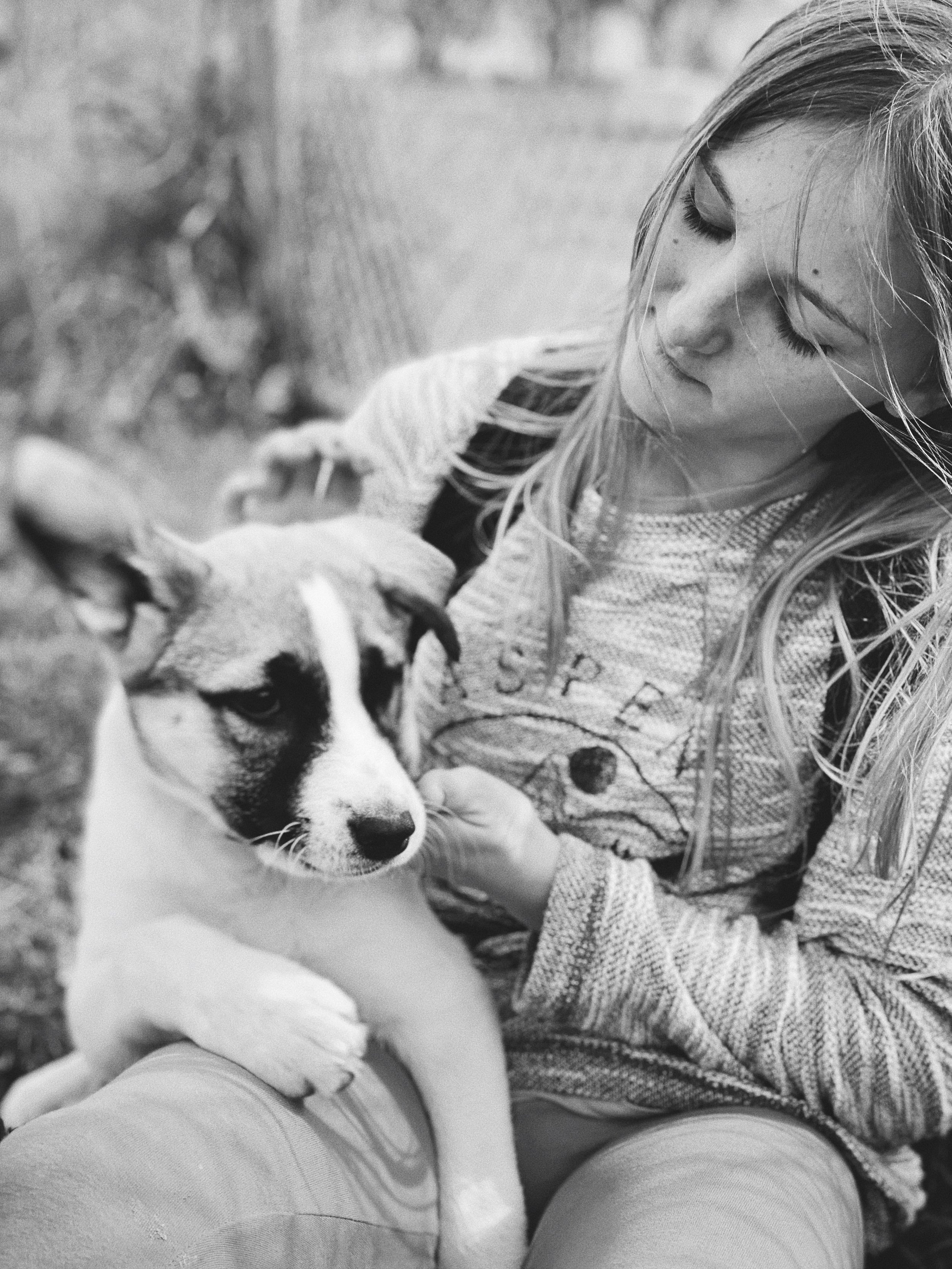 A young girl with long hair sitting outdoors with a puppy in her lap, holding its face and looking at it in black and white.