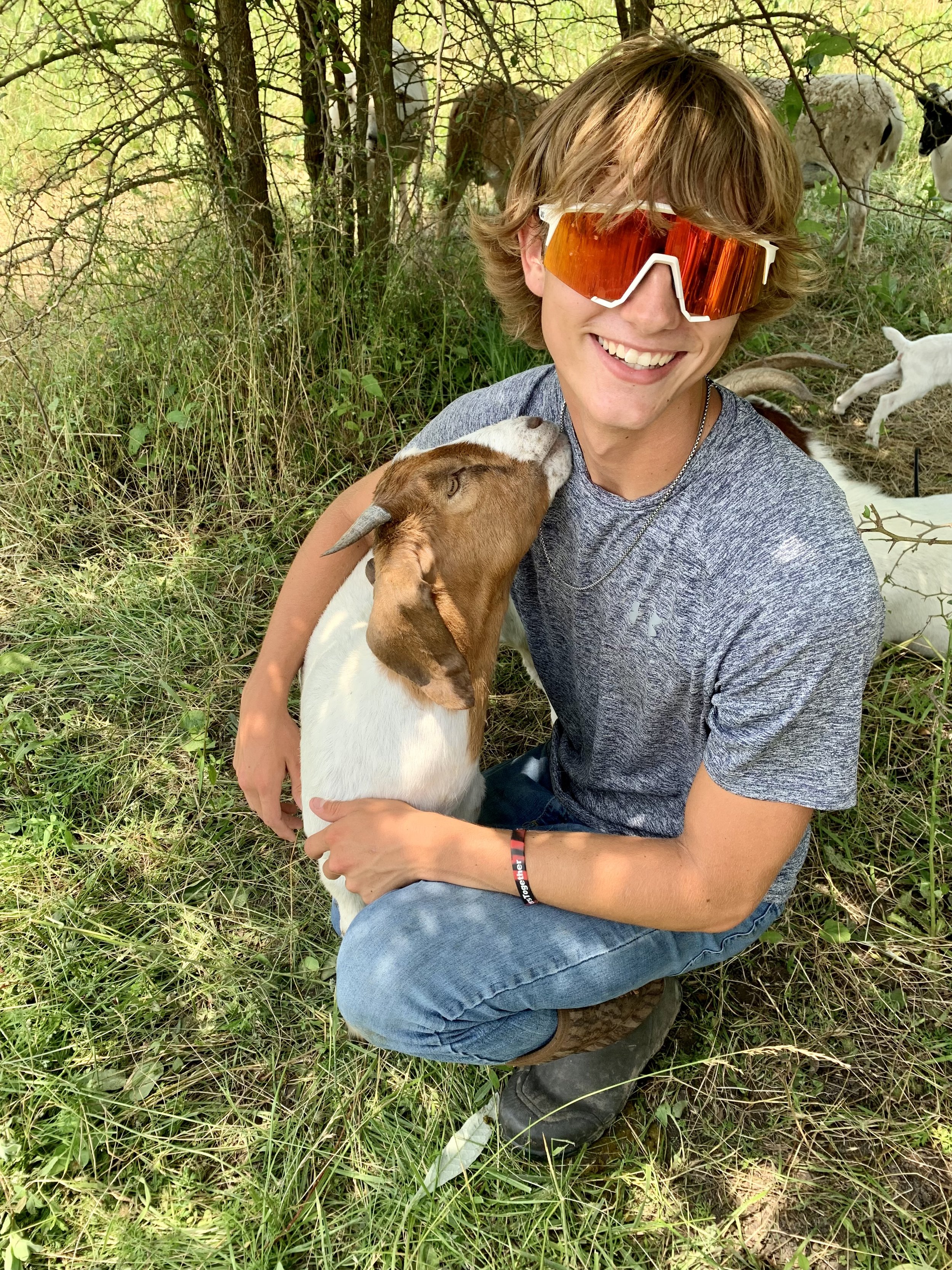 A young man with shoulder-length hair and orange-tinted sunglasses kneeling outdoors, smiling, and hugging a goat. The goat is licking his cheek. The background features green grass, trees, and other goats.