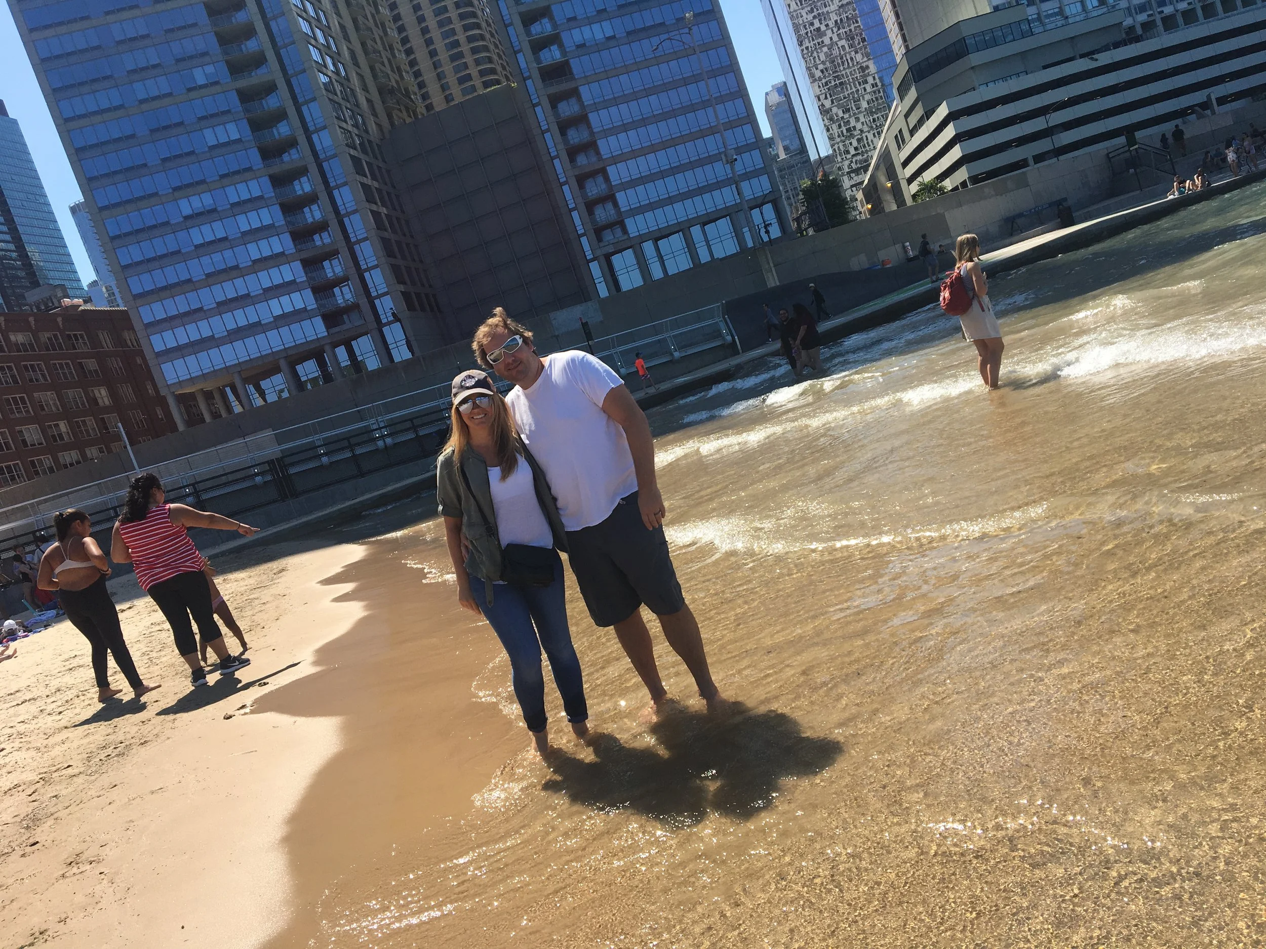 A couple standing in shallow water at a beach with tall city buildings in the background.