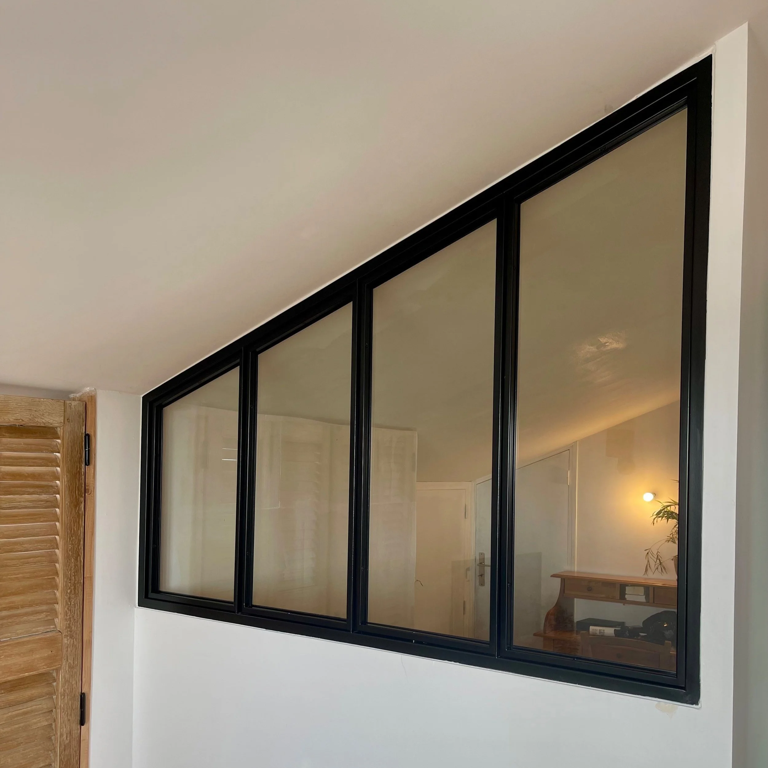 Fenêtre en verre avec cadre noir, située dans un mur blanc, avec vue sur un intérieur avec un meuble en bois, une plante et un éclairage chaud.