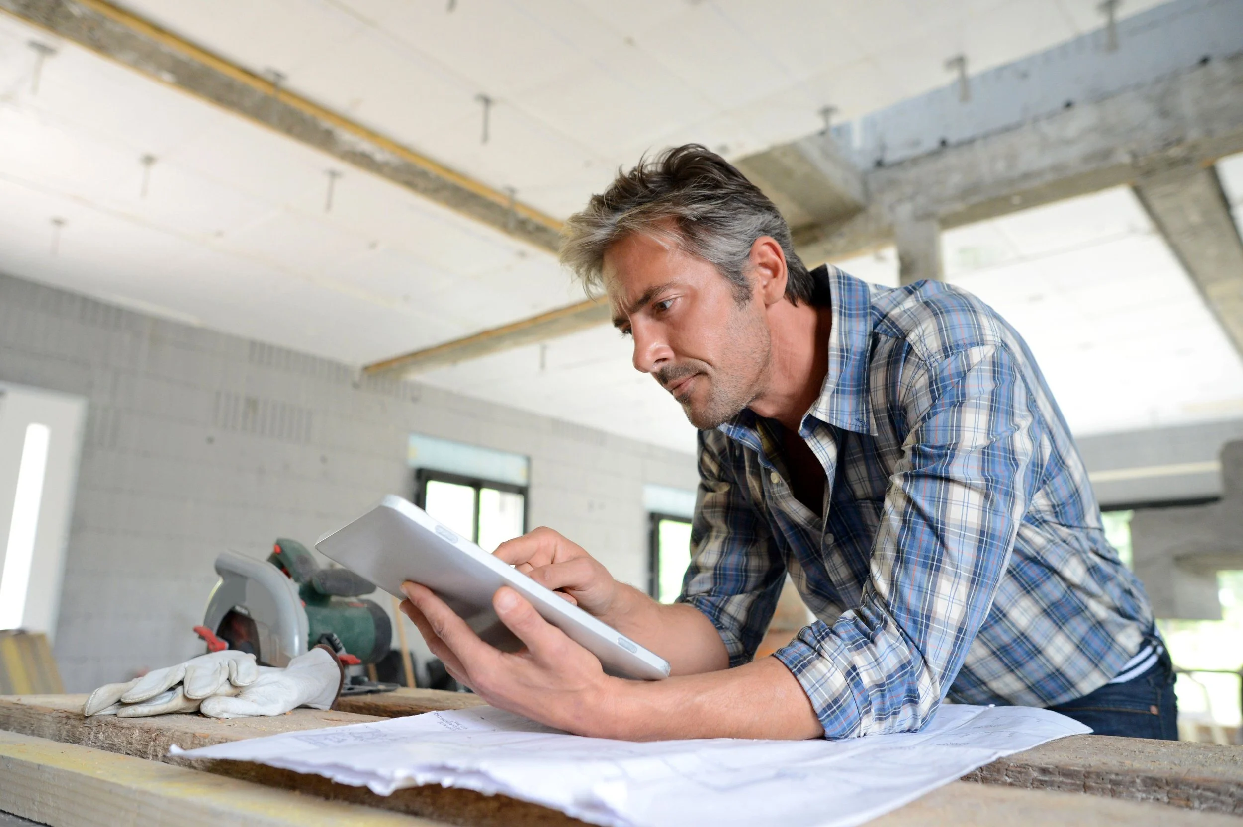 Construction worker in a plaid shirt reviewing project plans on a digital tablet at a job site, demonstrating the use of modern technology in construction management.