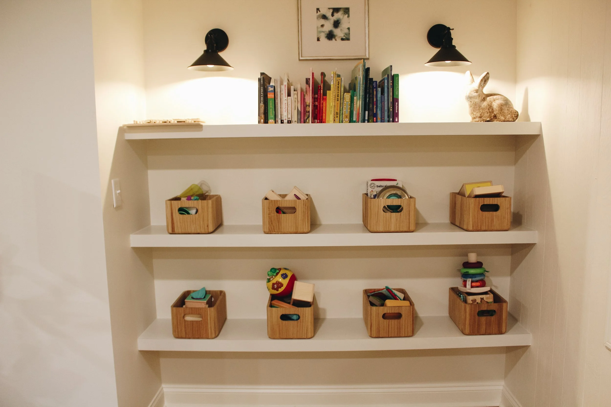 A built-in white bookshelf with three shelves, decorated with children's books, toys, and small wooden storage boxes filled with toys and art supplies. The top shelf has a framed picture and a rabbit figurine, with two wall-mounted black lamps illuminating the display.