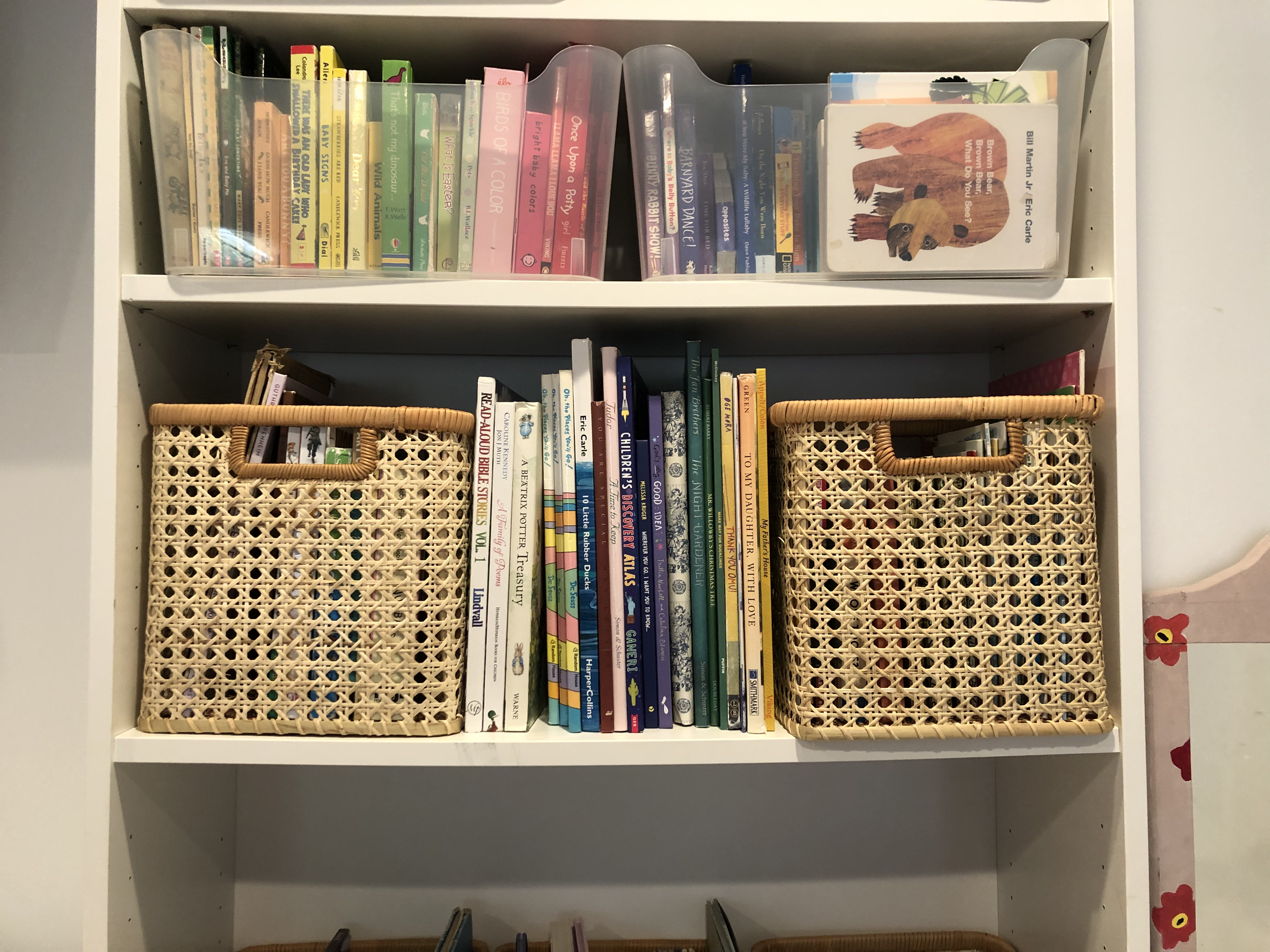 A white bookshelf with three shelves filled with children's books, some in transparent boxes, and two woven baskets containing books on the middle shelf.