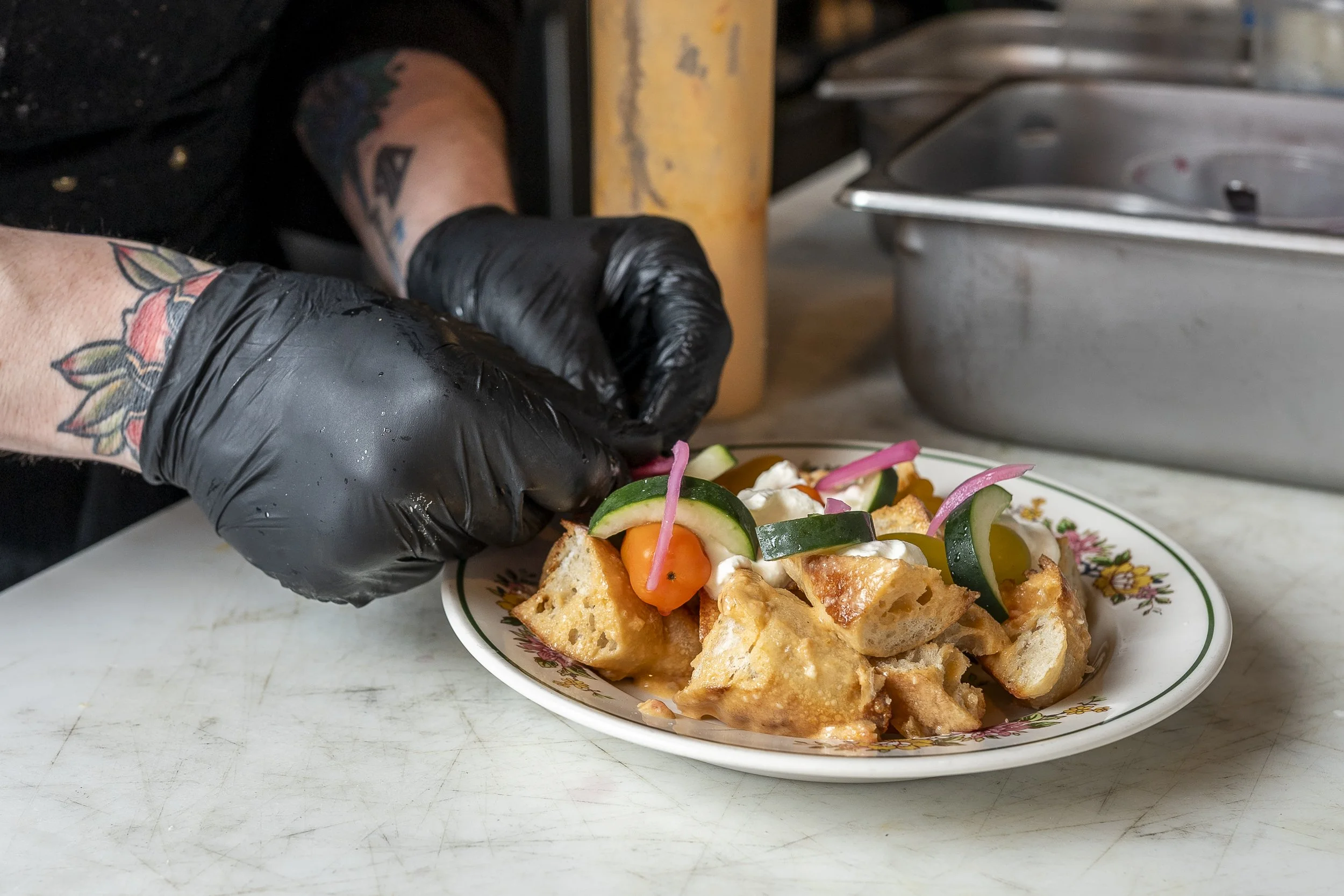 A person wearing black gloves and tattoos is preparing a salad with cucumbers, cherry tomatoes, pickled onions, and fried bread pieces in a floral bowl.