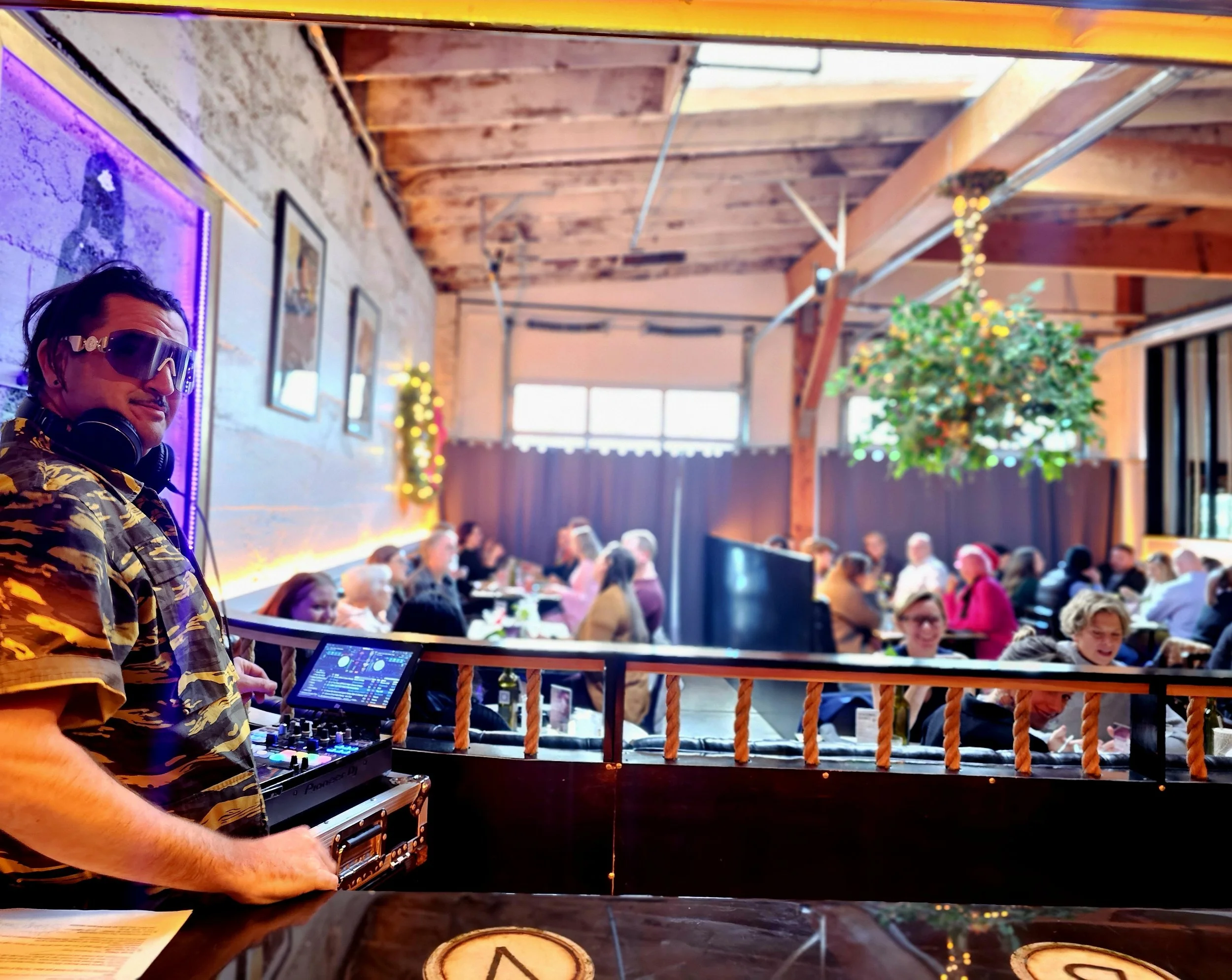 A man wearing a camo shirt and goggles is DJing at an indoor event with a large crowd. The room has a rustic wooden ceiling, string lights, and some greenery decorations.