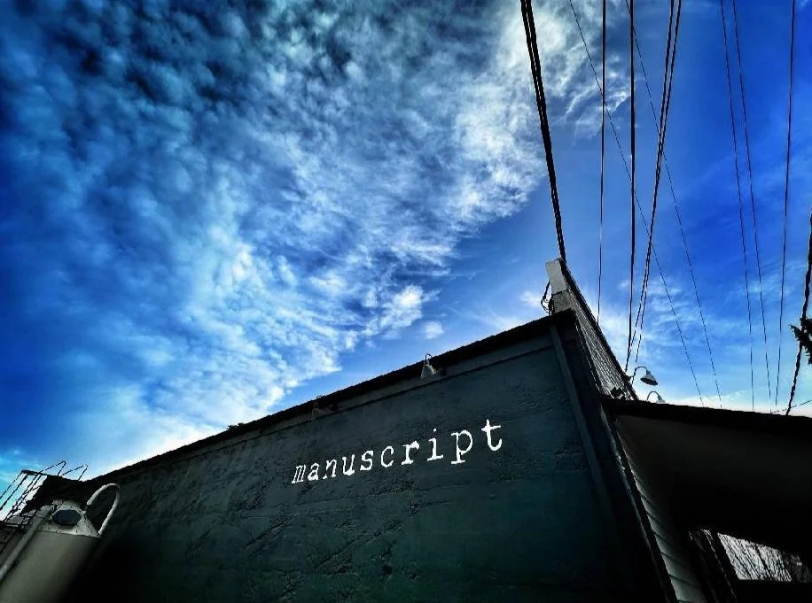 Outdoor shot of the exterior of the restaurant with the word 'manuscript' written on it, under a partly cloudy blue sky with power lines overhead.