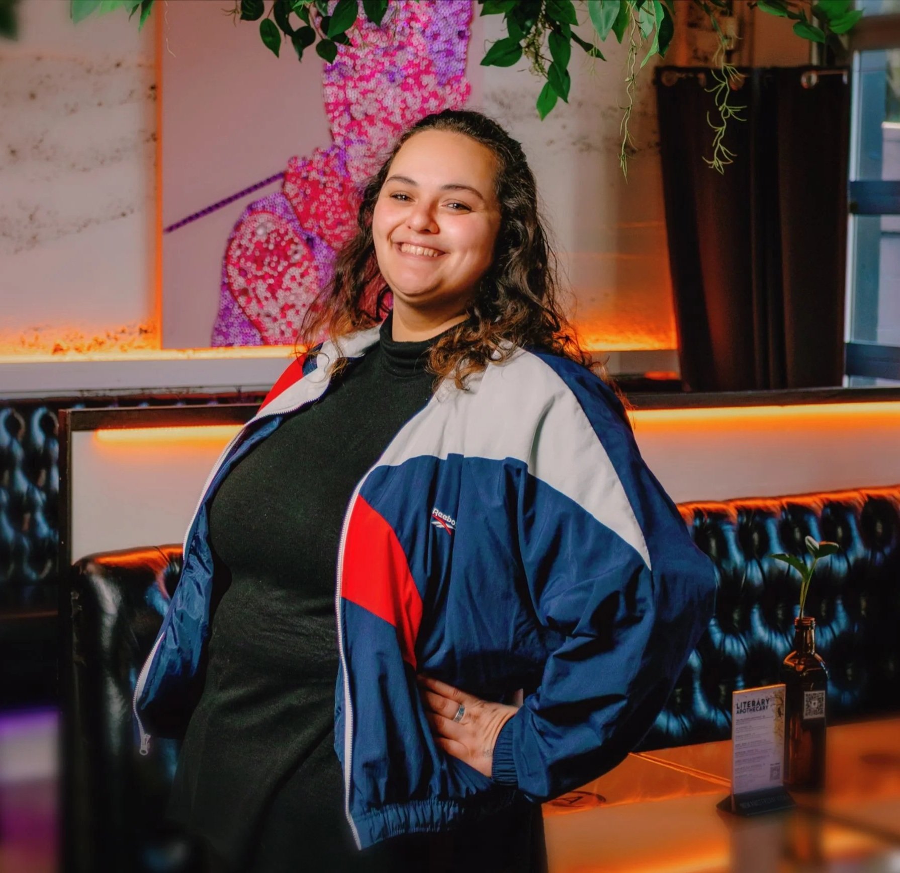 A smiling woman with curly hair wearing a black top and a blue, white, and red jacket in an indoor environment with warm lighting. Behind her, there is a wall with a colorful pink, purple, and red art piece, hanging plants, and part of a dark wooden curtain. There is a black leather chair and a display stand with a bottle and informational card on the right.