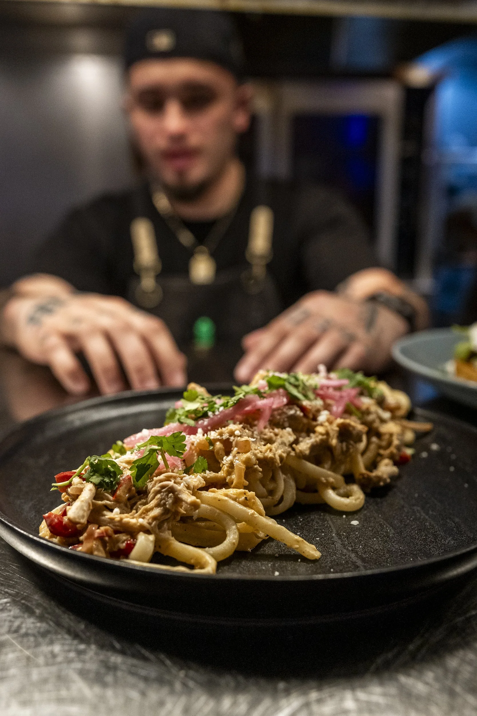 Close-up of a plate of pasta with vegetables and toppings, while a chef in black clothing is working in the background.