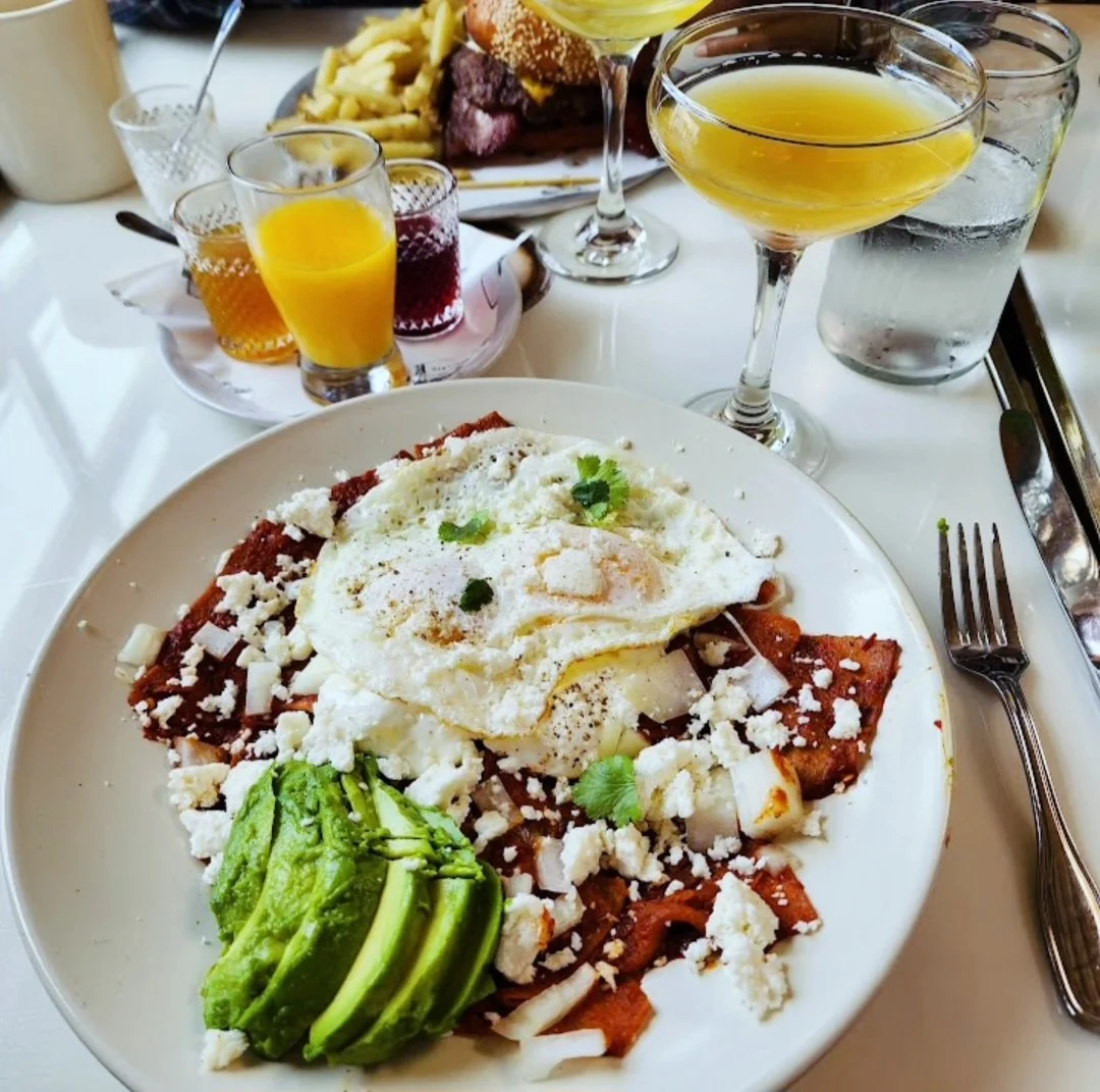 A plate of Mexican chilaquiles with a fried egg, avocado slices, and crumbled cheese, surrounded by drinks including orange juice and cocktails, on a white table.