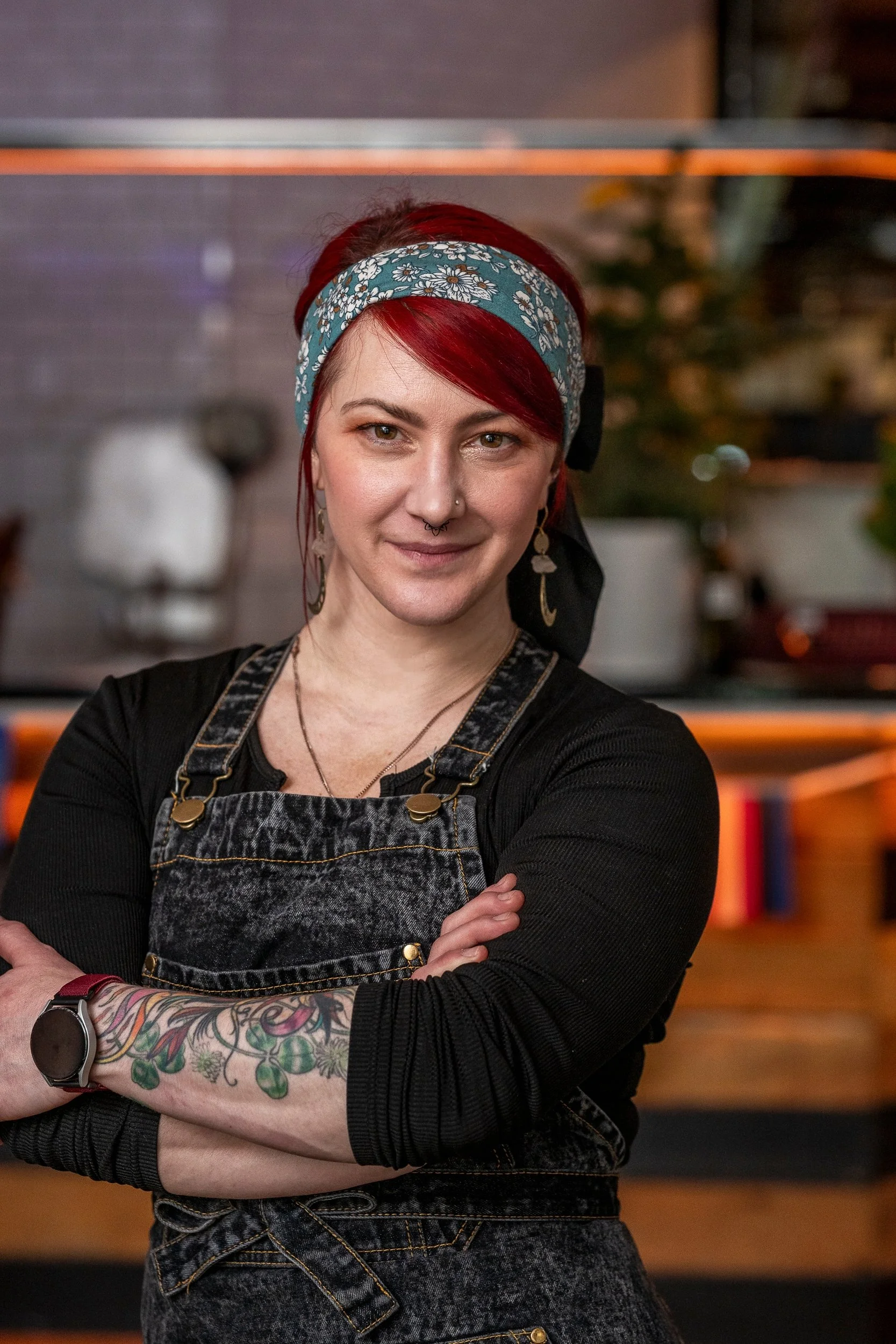 A woman with red hair, wearing a blue floral headband and a black long-sleeved shirt, standing indoors with her arms crossed, smiling slightly, with books and potted plants in the background.