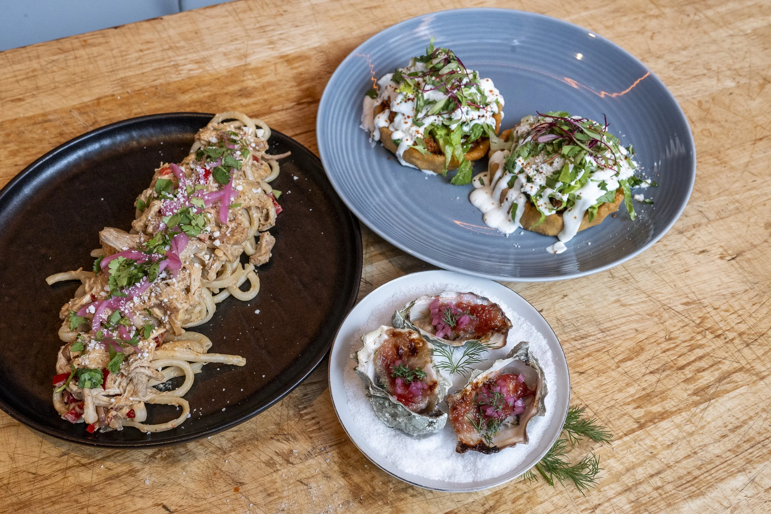 A variety of tacos and pasta dishes on a wooden table, featuring topped tacos with greens and sauces, and a pasta with chicken, vegetables, and herbs.