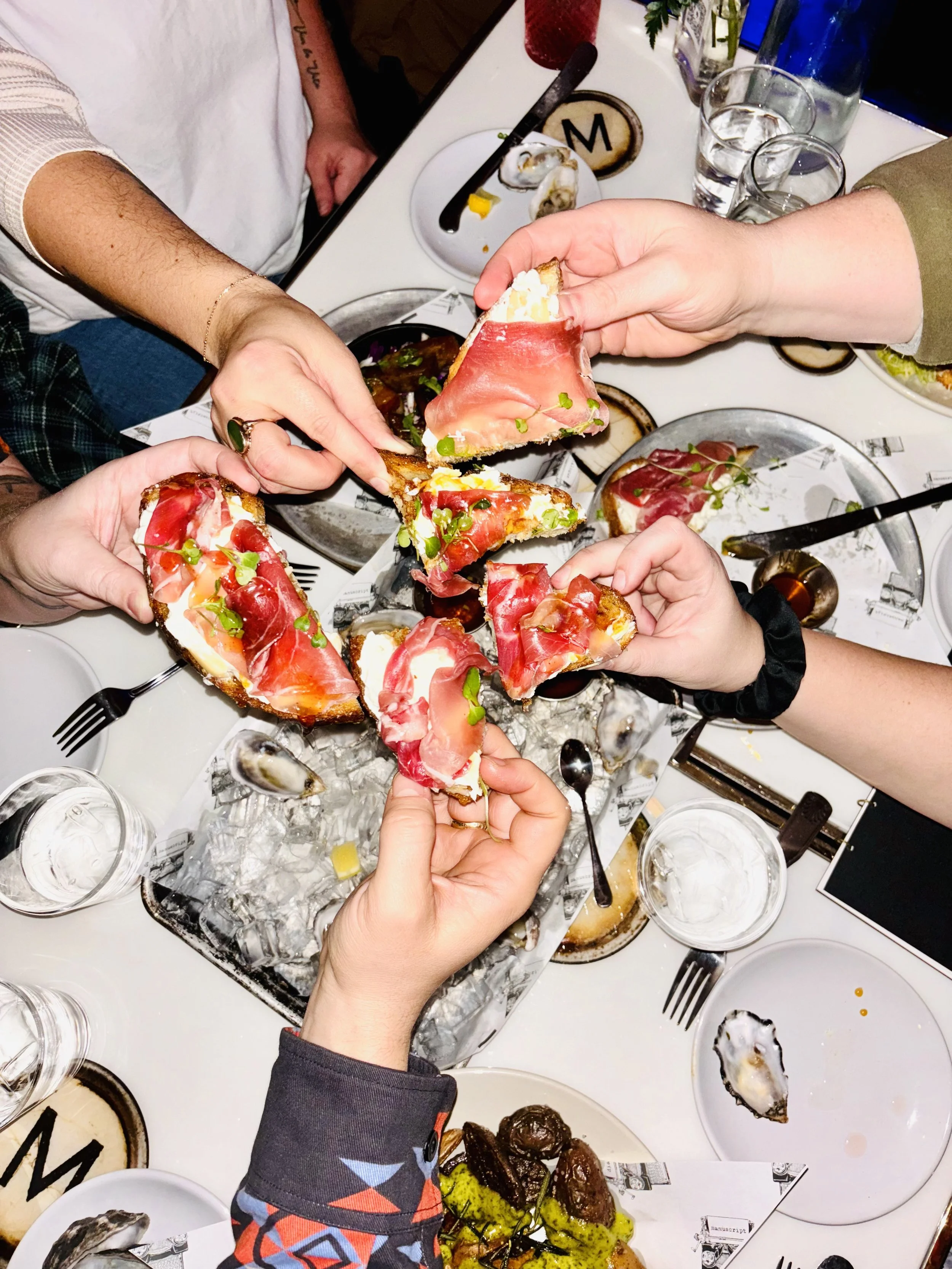 People sharing slices of pizza topped with prosciutto and microgreens at a table with drinks and various plates of food.