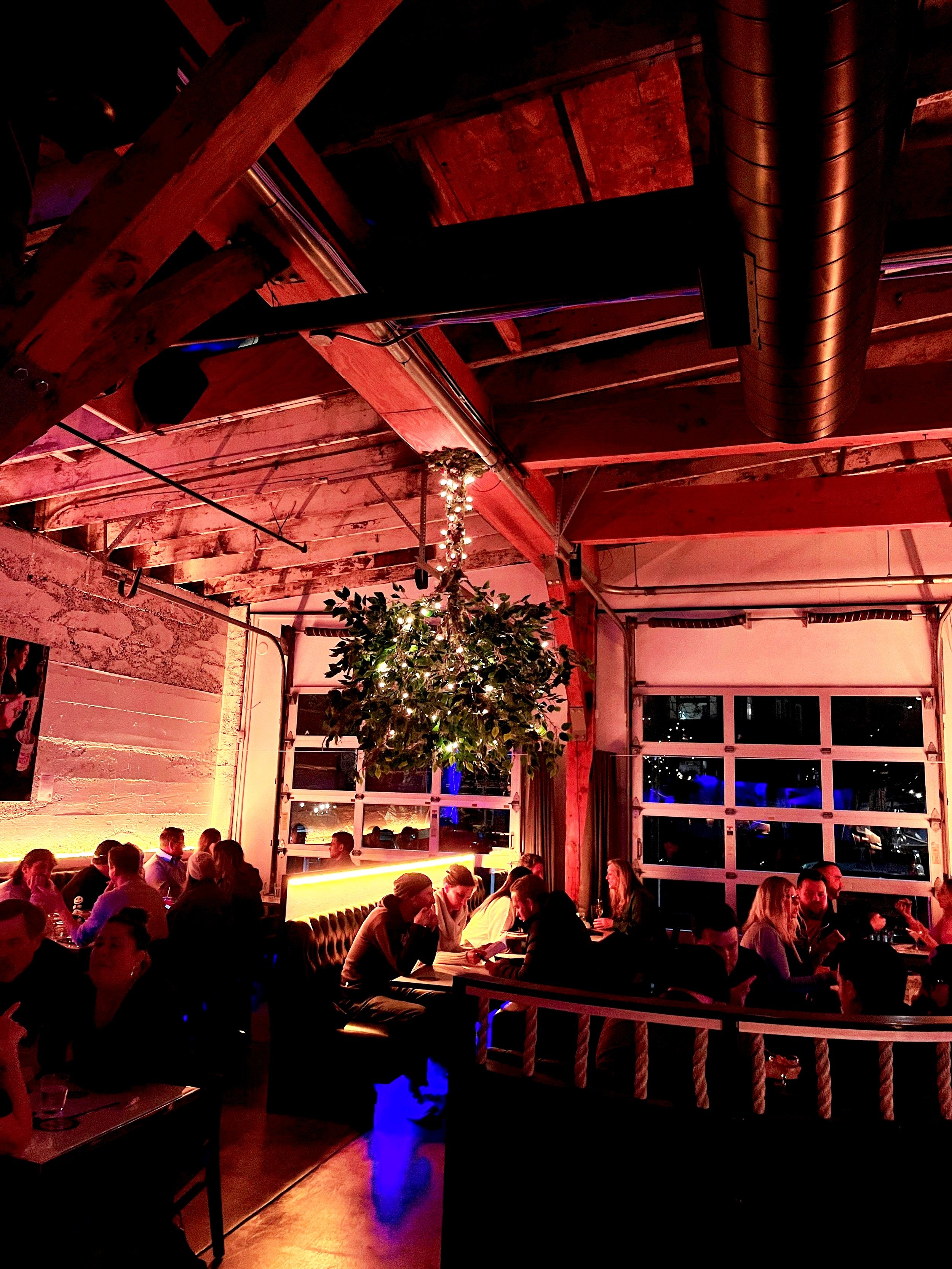 People dining in a warmly lit restaurant with exposed brick walls, a large window, and a decorated ceiling with plants and string lights.